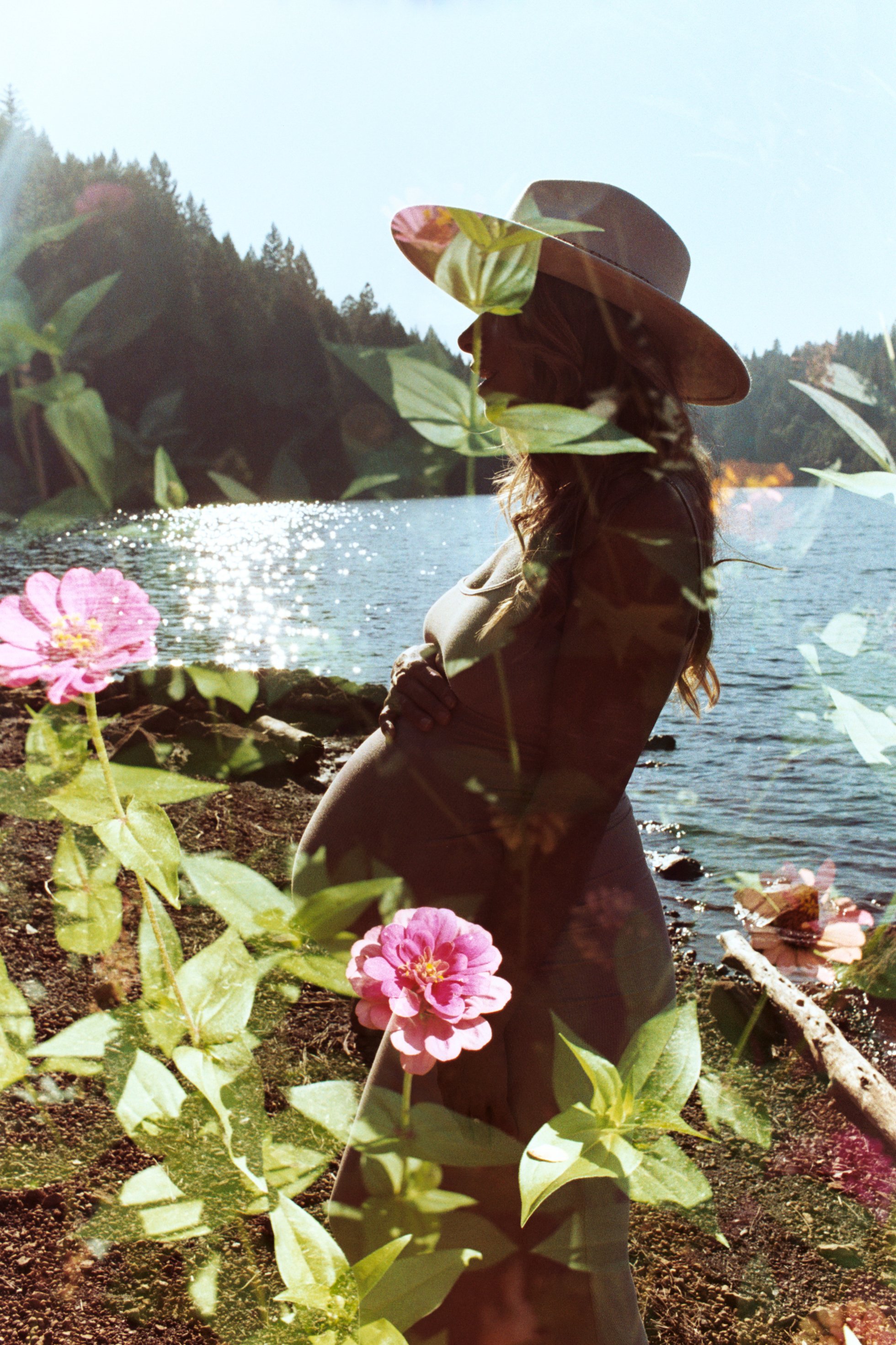 A woman with long hair wearing a wide-brimmed hat standing by a lake with mountains in the background, surrounded by pink flowers and green foliage.