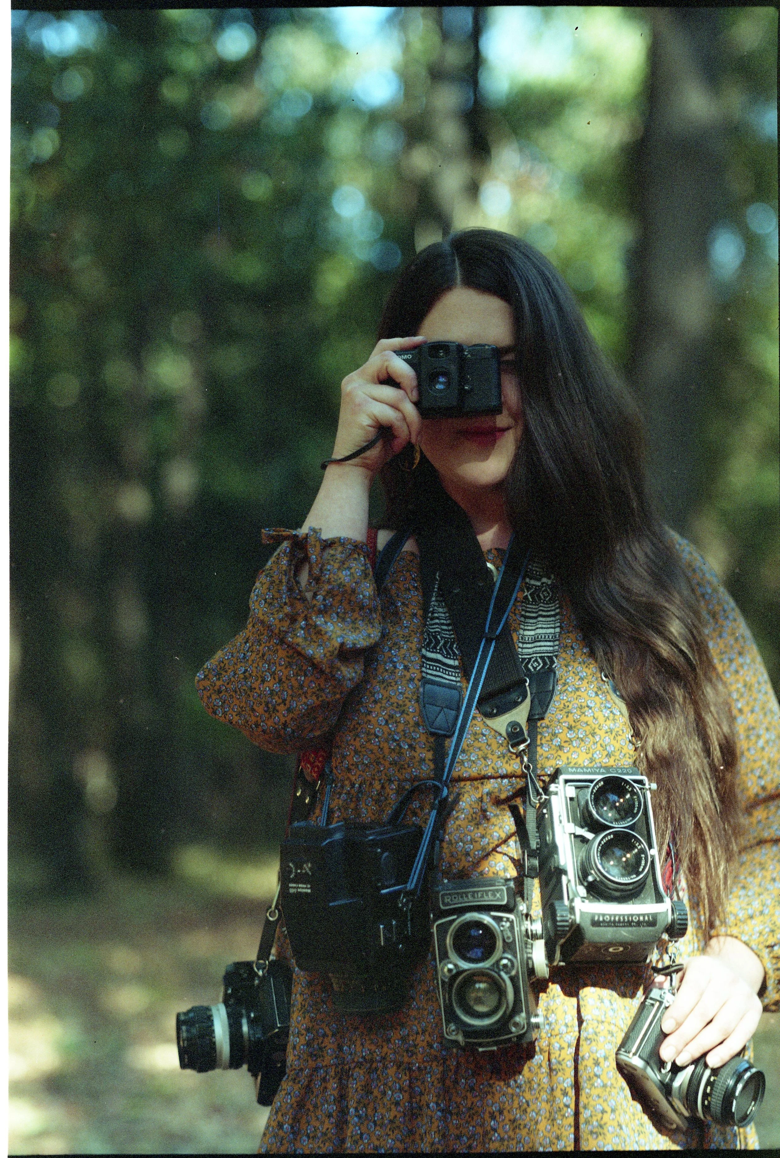 A woman with long, wavy hair wearing a patterned dress is holding a camera up to her face, taking a photo outdoors in a green, wooded area. Multiple vintage cameras are hanging around her neck and waist.