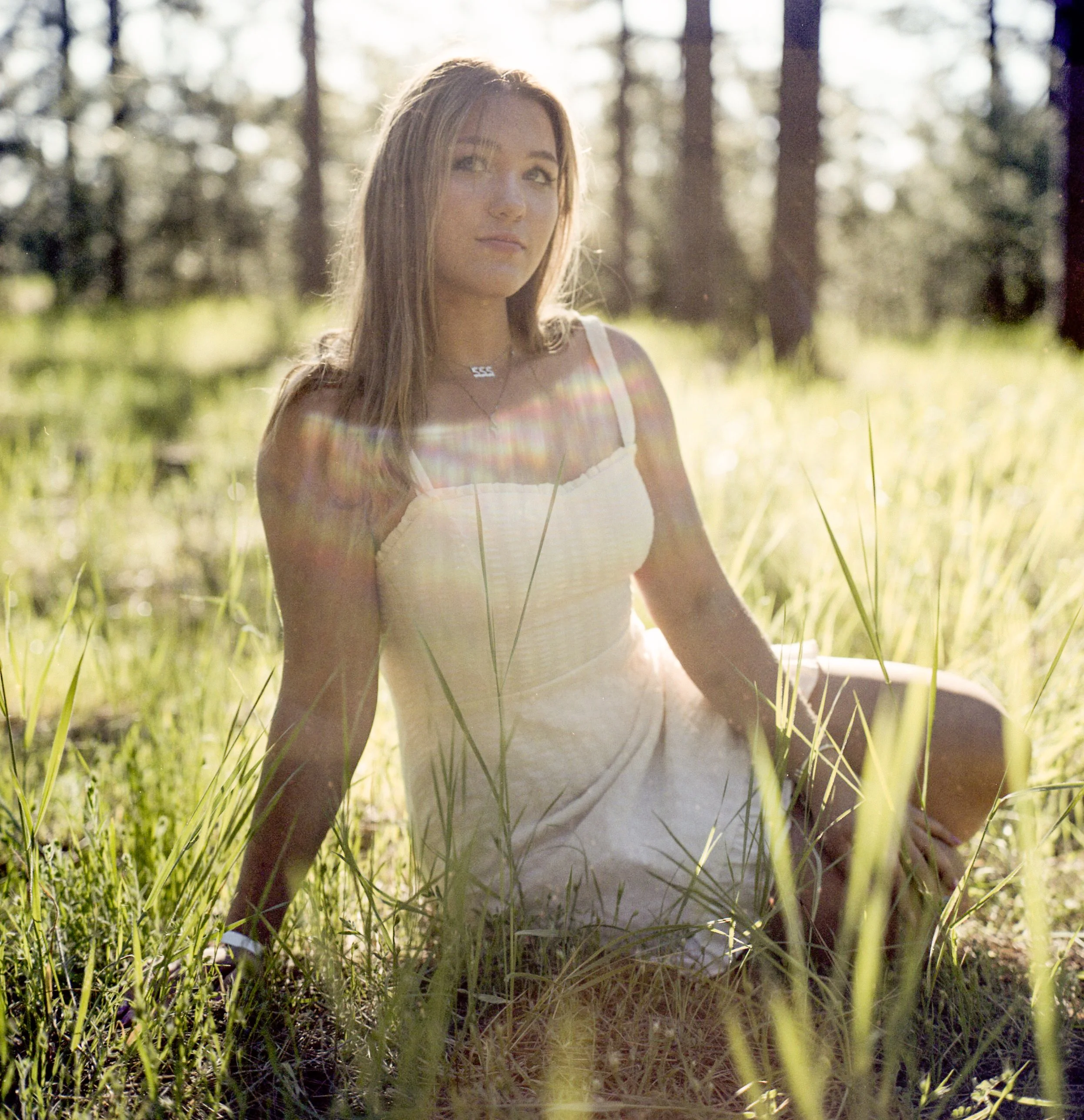 A young woman with long hair sitting in a grassy field with trees in the background, backlit by sunlight.