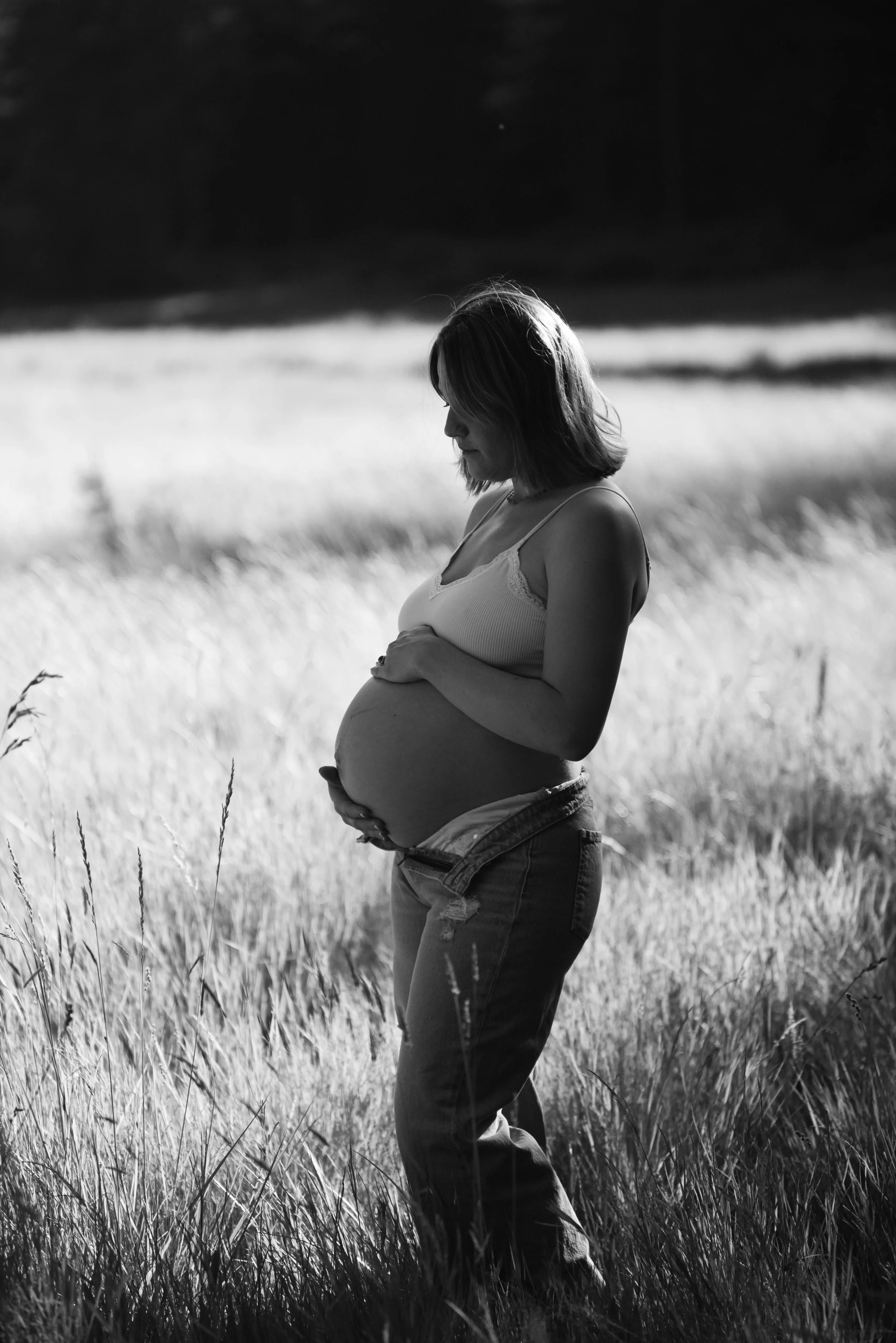 A pregnant woman standing in a field, looking down and holding her belly.