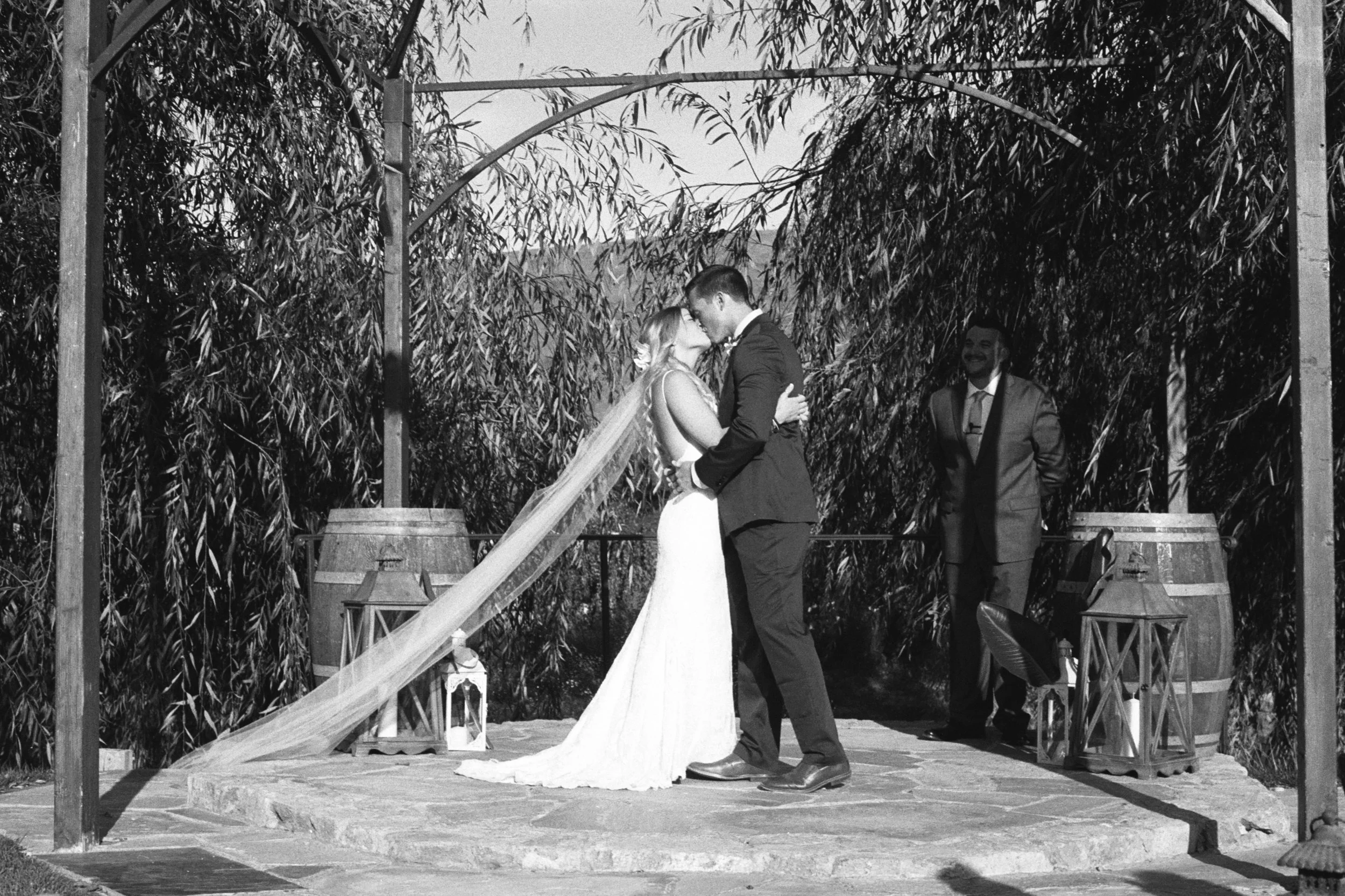 A bride and groom sharing a kiss during their outdoor wedding ceremony, with a man in a suit off to the side, and trees and a wooden structure in the background.