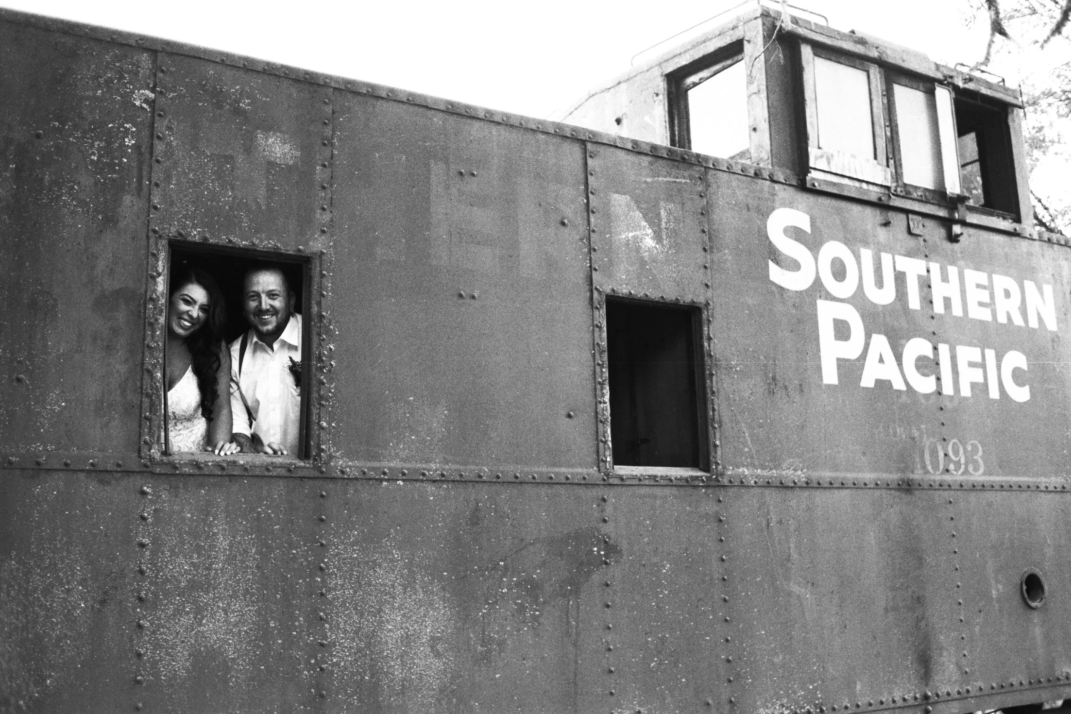 Black-and-white photo of a rusty train car with two windows. A couple, a woman and a man, are looking out of the lower window, smiling. The train car has the words 'SOUTHERN PACIFIC' on its side.