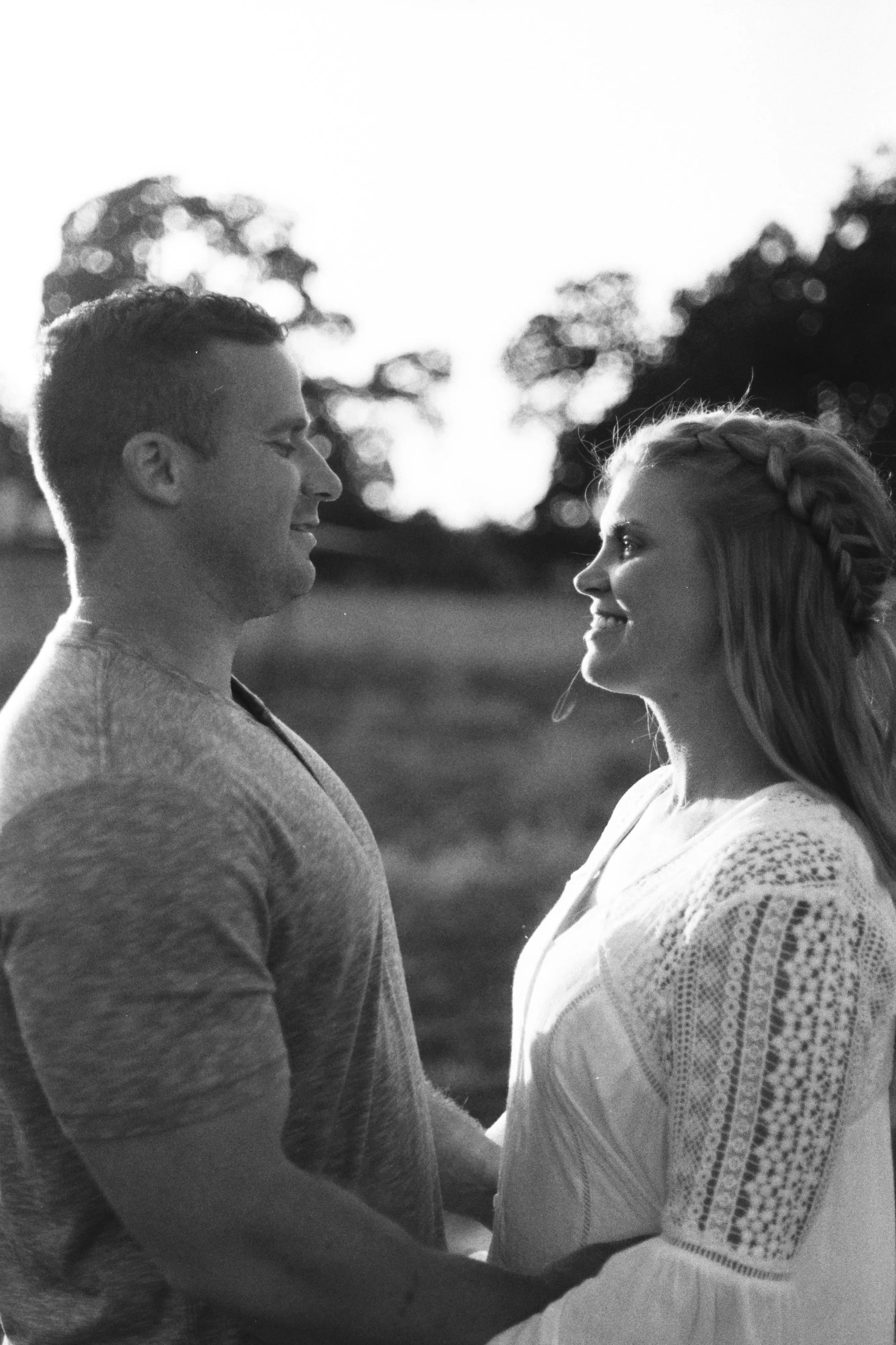 A black and white photo of a man and a woman facing each other, smiling, outdoors with blurred trees in the background.