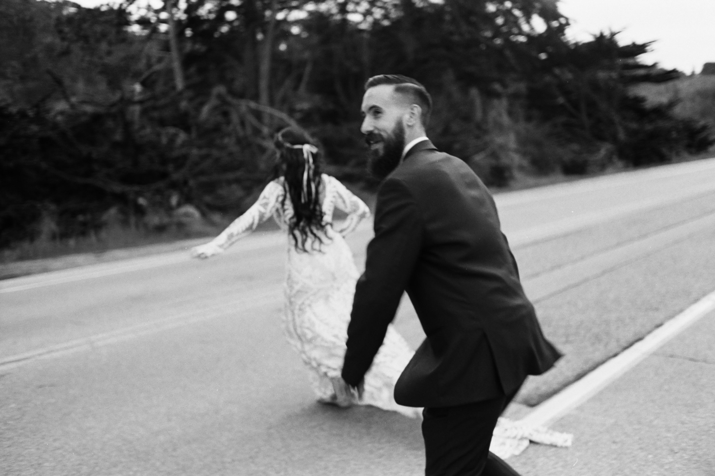 A man in a suit with a beard and hairstyle, walking on the side of a road with trees and bushes in the background during daytime