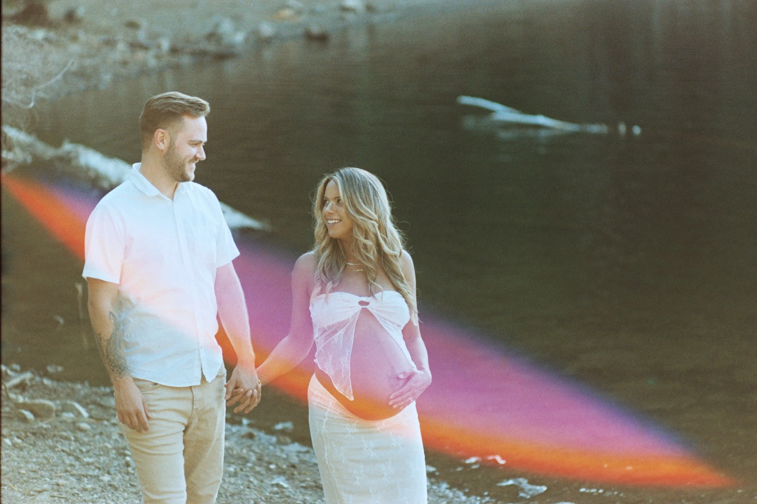 A man and woman holding hands, walking on a rocky beach near the water, with the woman appearing pregnant and both smiling at each other.