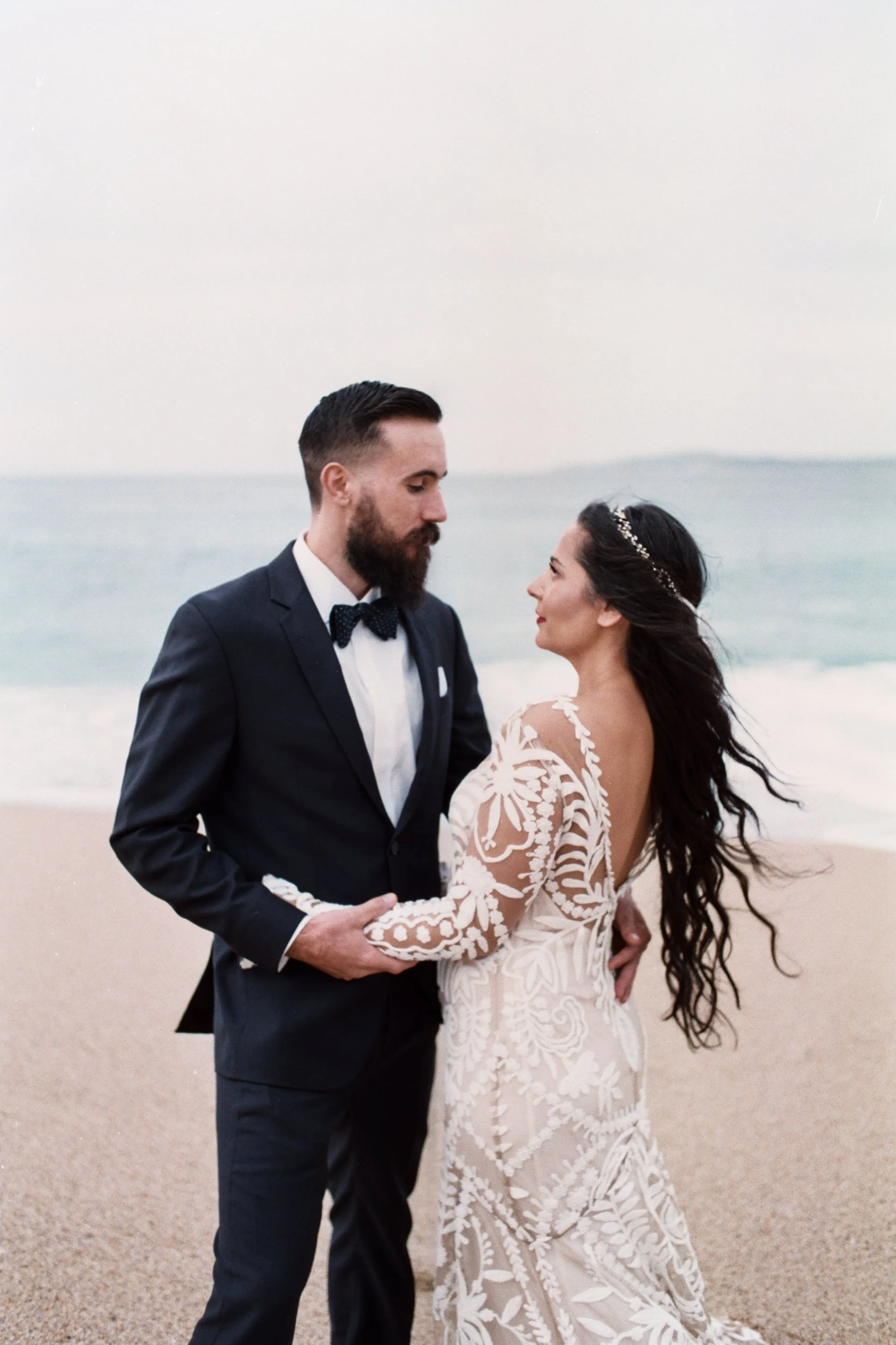 A bride and groom stand close together on a beach, holding hands and looking at each other, with the ocean in the background.