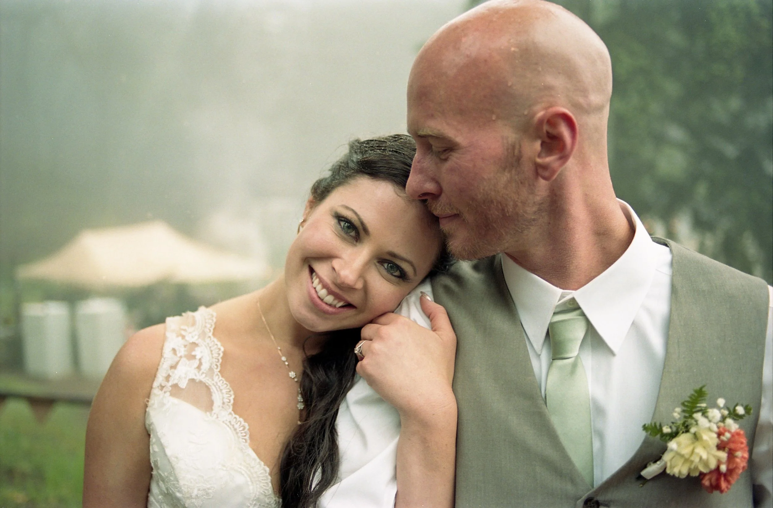 A bride and groom share an intimate moment, with the bride smiling and resting her head on the groom's forehead, in a lush outdoor setting.