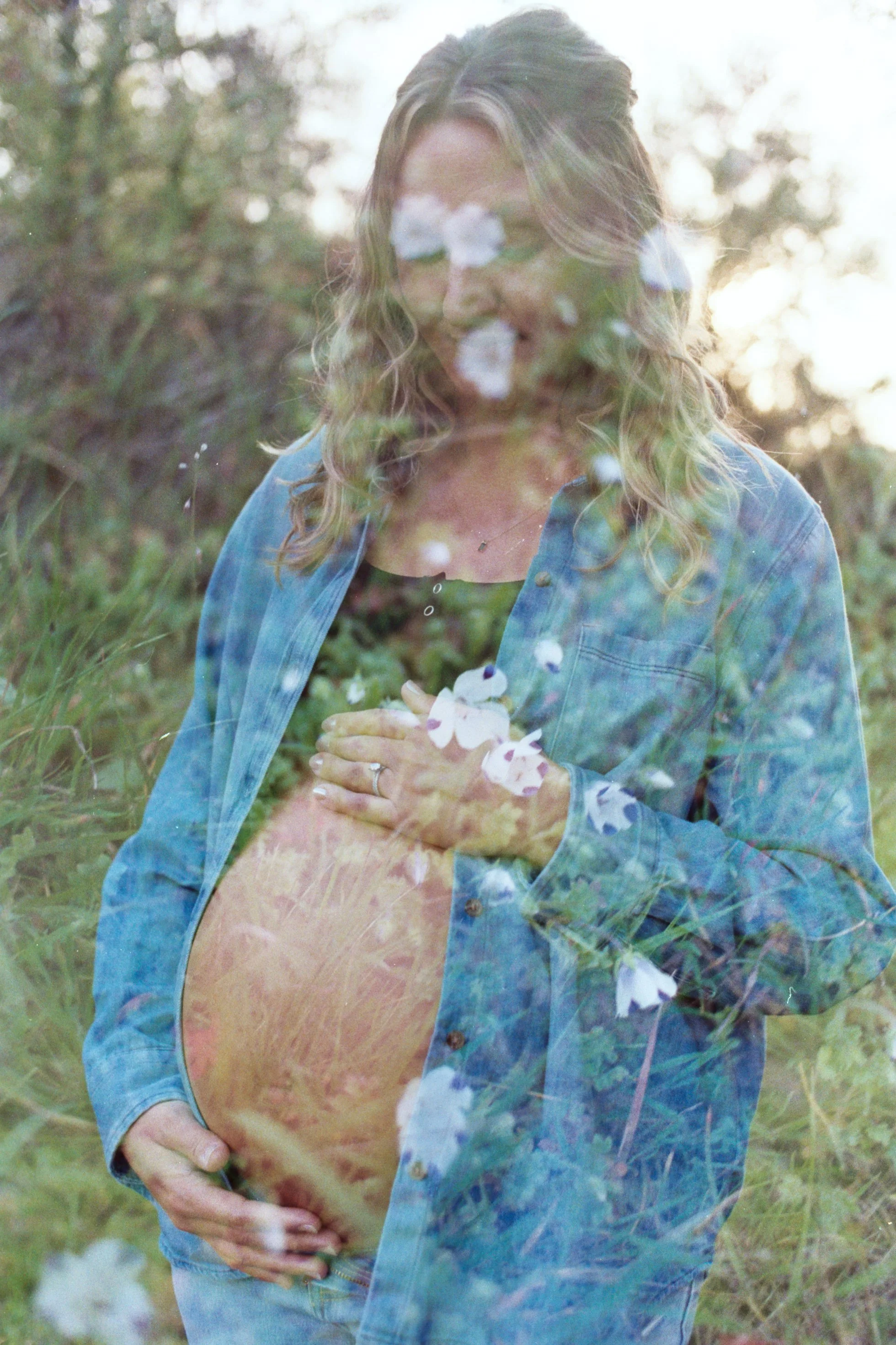 A woman is standing outdoors in a grassy area with trees, smiling with her eyes closed. She has wavy hair and is wearing a denim jacket over her belly, which suggests she is pregnant, and she is cradling her belly with her right hand. The photo has a