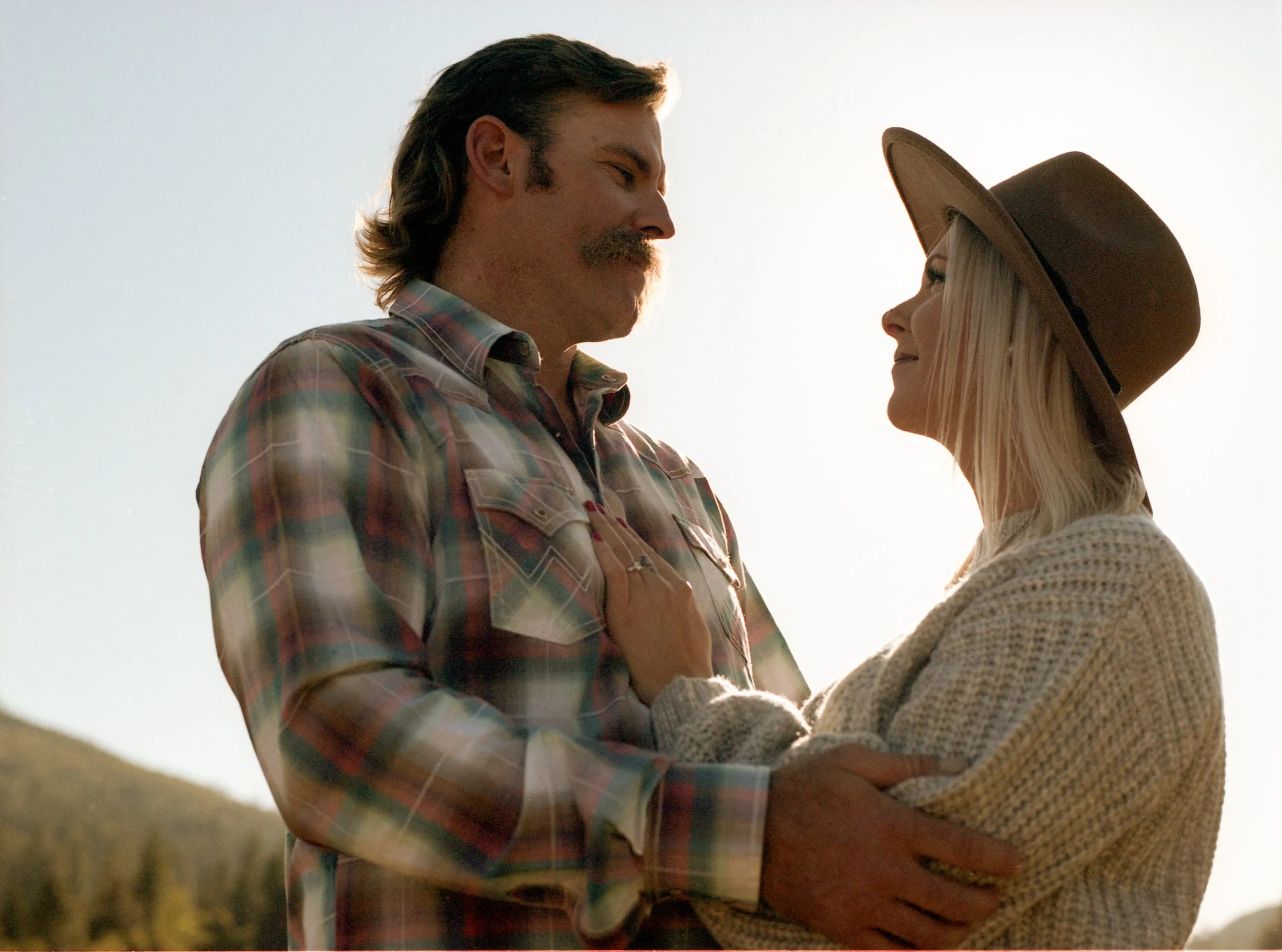 A man and woman look at each other during sunset or sunrise, with the man wearing a plaid shirt and the woman wearing a wide-brimmed hat and sweater.