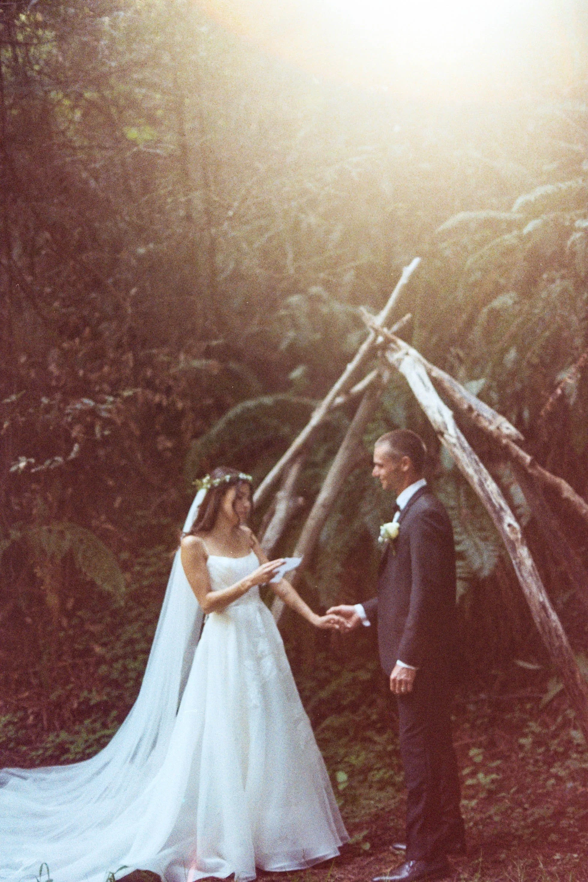 A bride and groom exchanging vows outdoors during a wedding ceremony, standing beneath a makeshift wooden arch with a background of trees and sunlight.