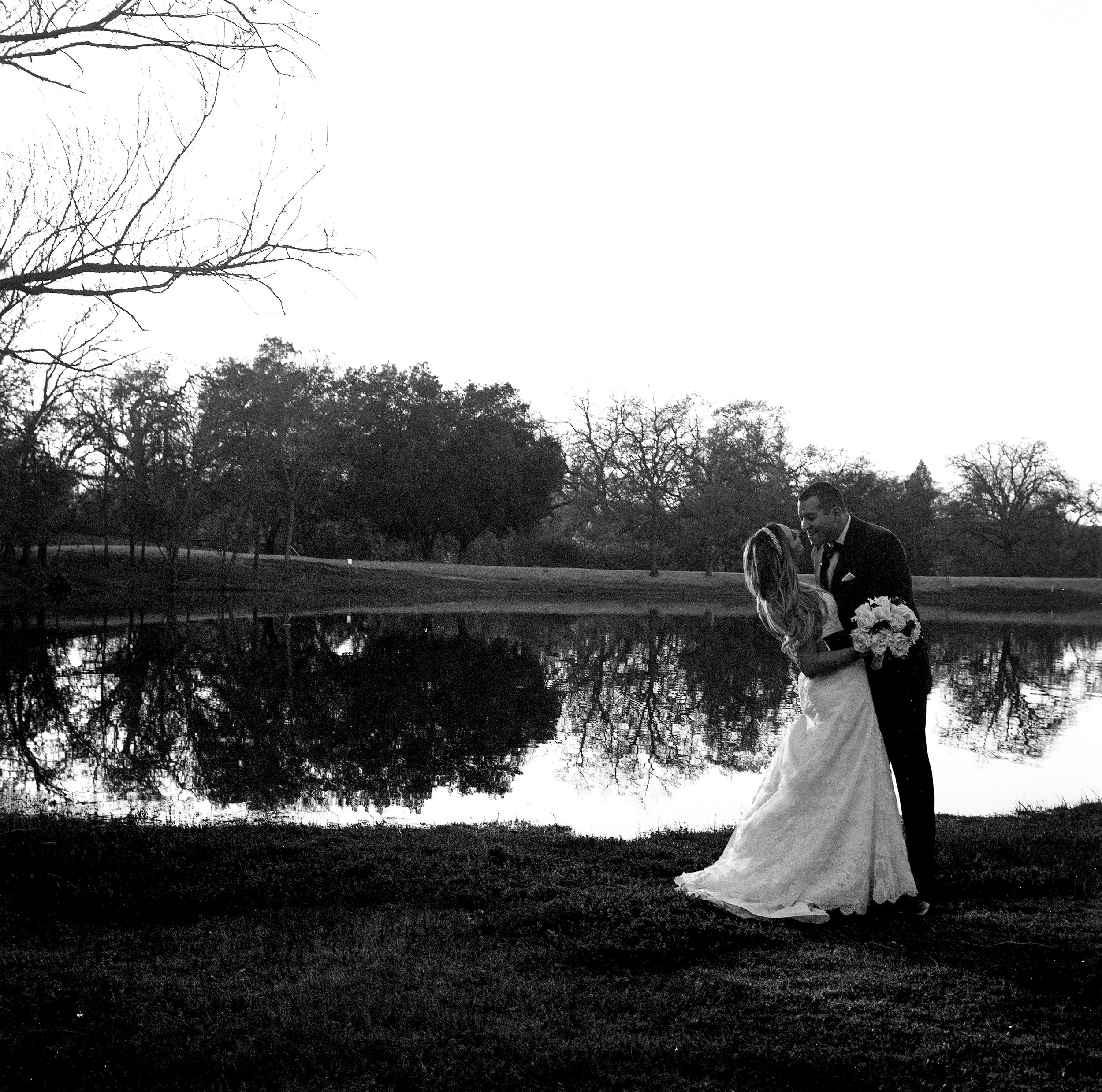 A black and white photo of a couple dressed in formal attire, standing by a lake with trees in the background. The woman is holding a bouquet of flowers, and they are engaged in a moment of affection.