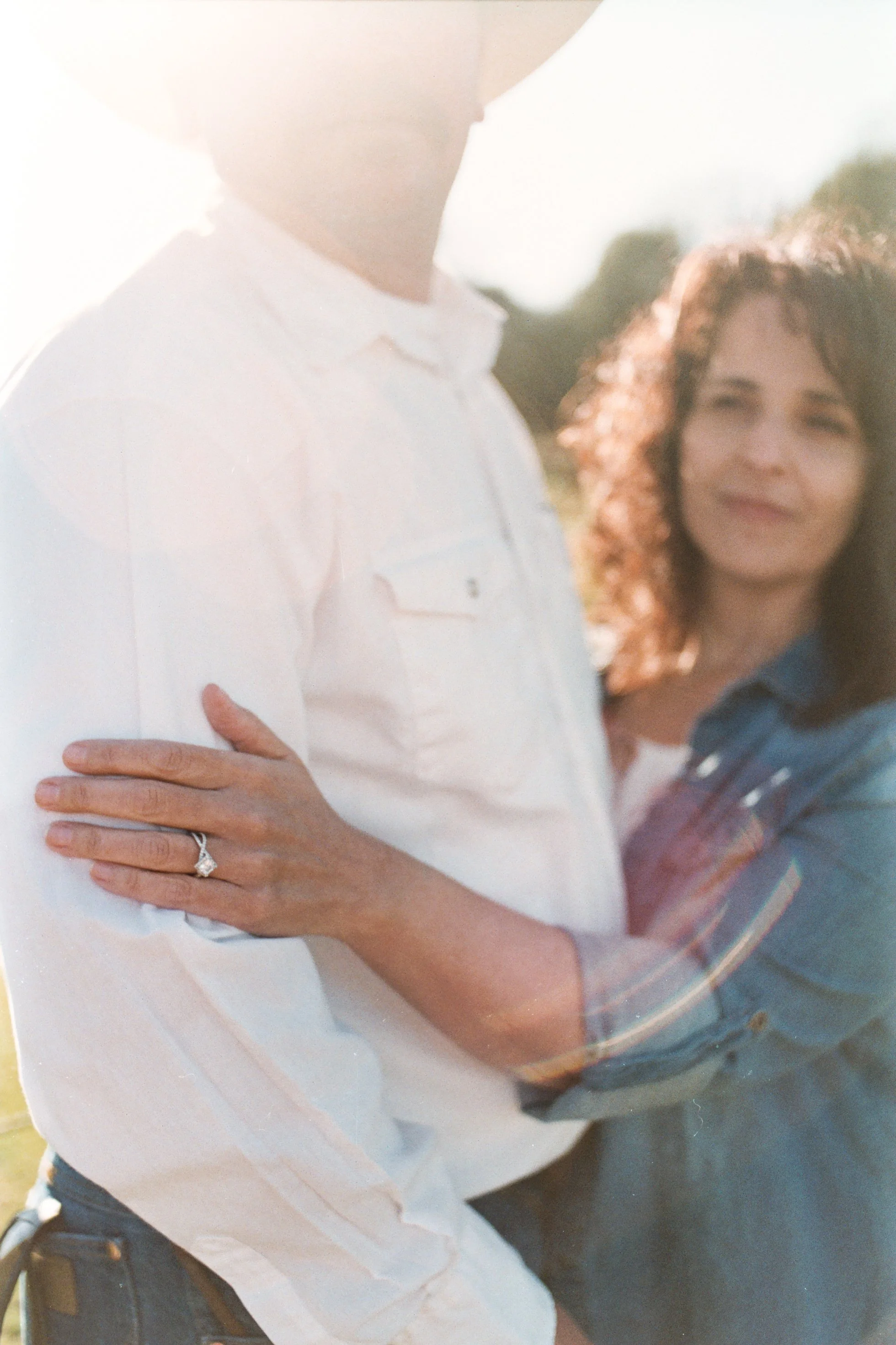 Close-up of a woman with curly hair smiling, wearing a denim shirt, with a partial view of a man in a white shirt. The woman's hand is on the man's chest, showcasing an engagement ring. The background is outdoors with bright sunlight.