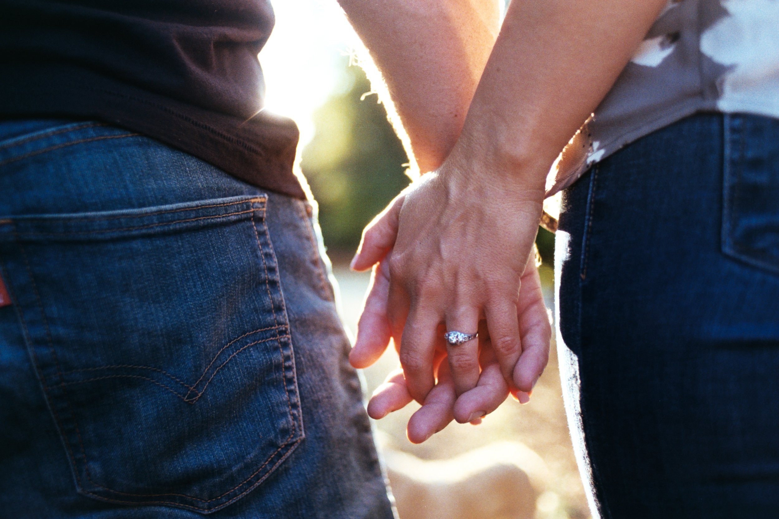Close-up of a couple holding hands, with the woman wearing a diamond ring and an engagement ring, during a sunset or sunrise.