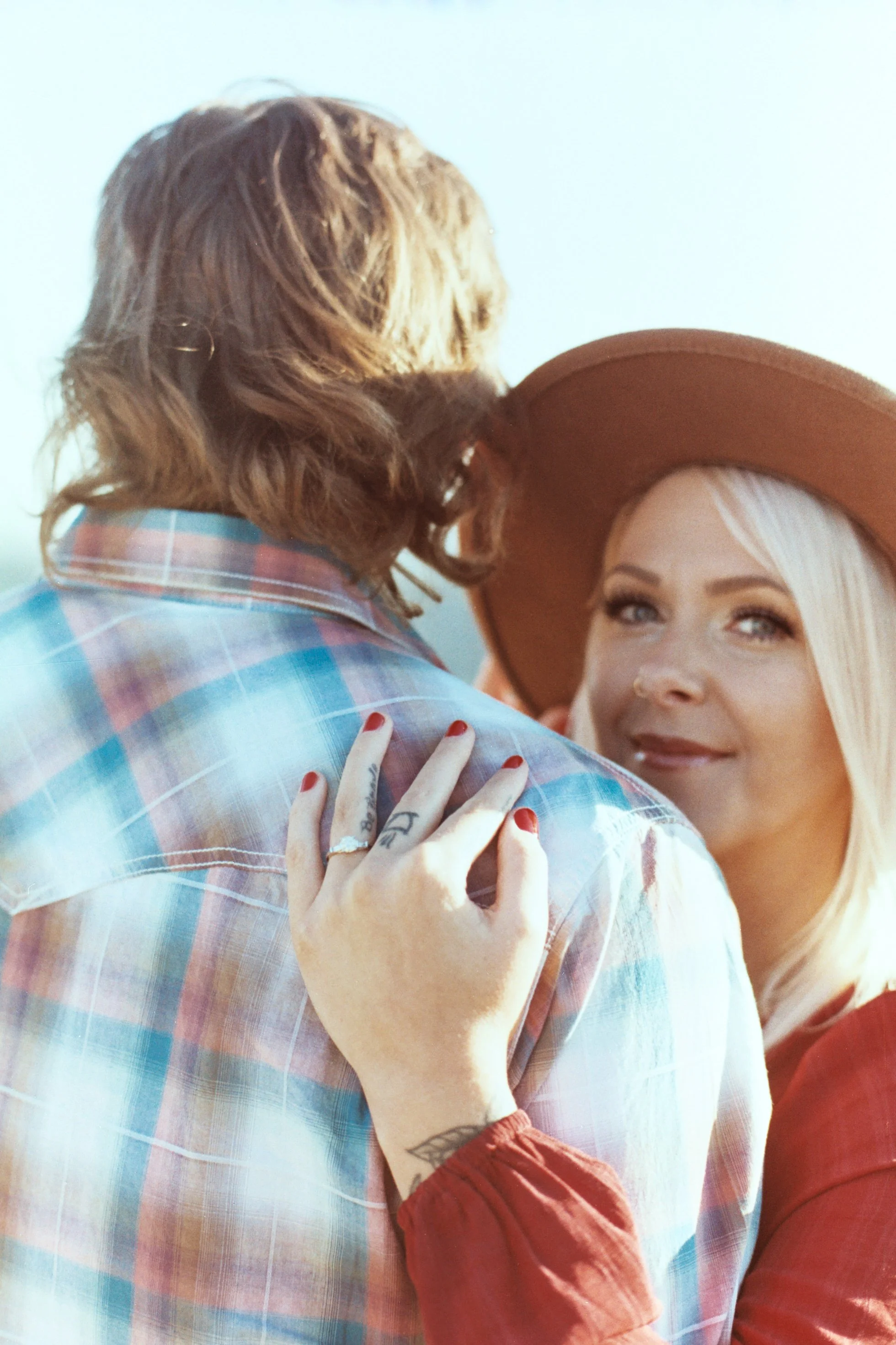 A man and a woman are embracing. The woman's face is visible, and she is wearing a wide-brimmed hat and has blonde hair and blue eyes. The man has long, curly hair and is wearing a plaid shirt.