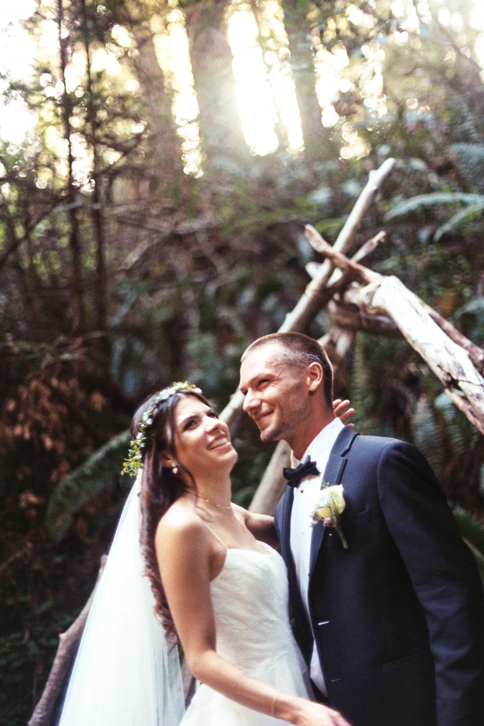 A bride and groom smiling and looking at each other during their wedding ceremony in a forest with sunlight shining through the trees, the bride wearing a white gown and a floral crown, and the groom in a black tuxedo with a bow tie.