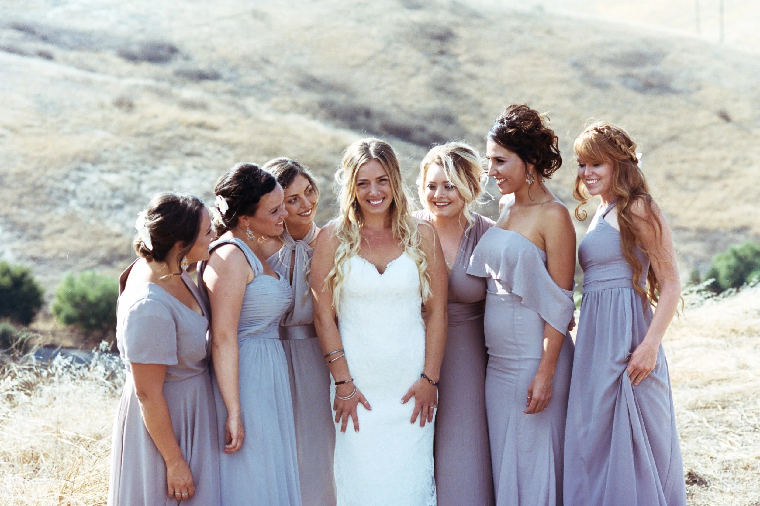 A group of women, including a bride in a white dress, standing outdoors on a dry, grassy landscape, with hills in the background, smiling and talking.