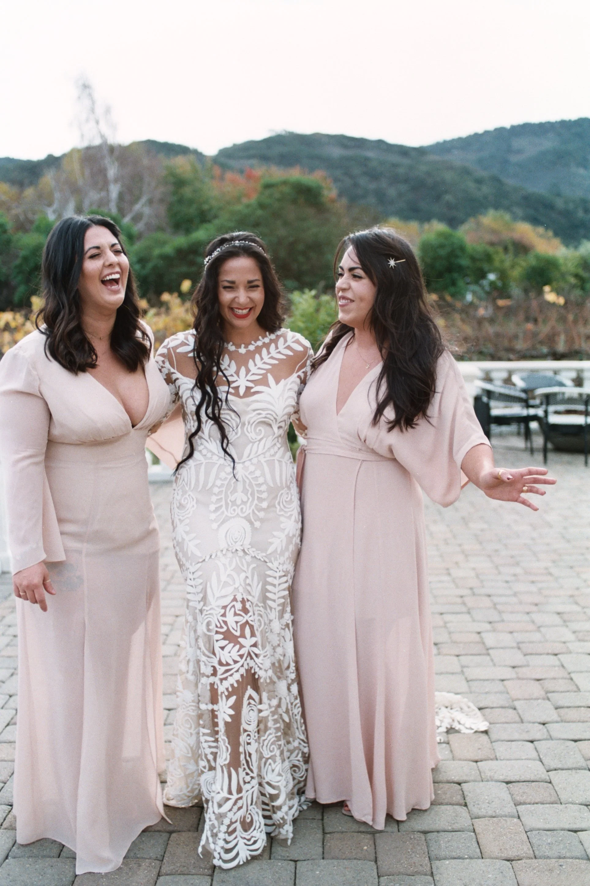 Three women dressed in light pink dresses standing outdoors on a cobblestone path with a scenic background of hills and trees, smiling and laughing together.