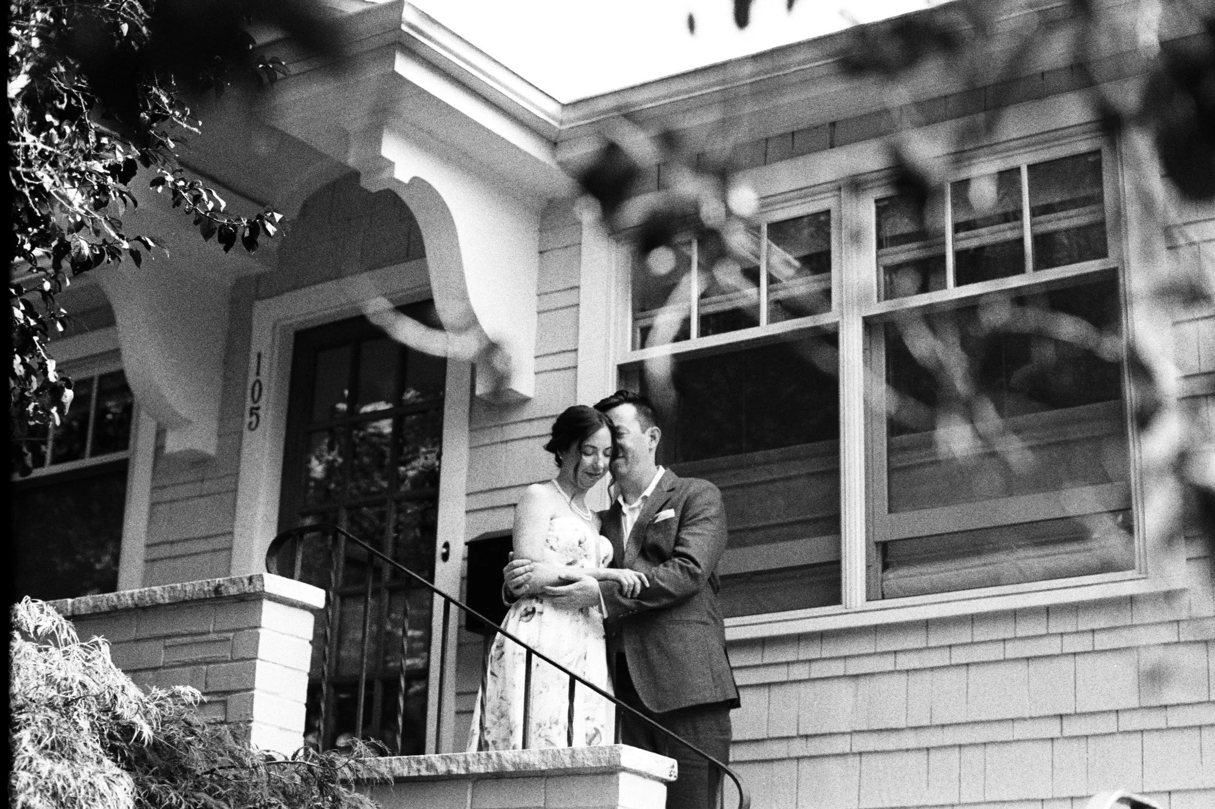 A black and white photo of a couple standing close together on a porch of a house, sharing an intimate moment. The woman has curly hair and is wearing a dress, the man is in a suit. The house has siding, large windows, and a decorative front door. Th