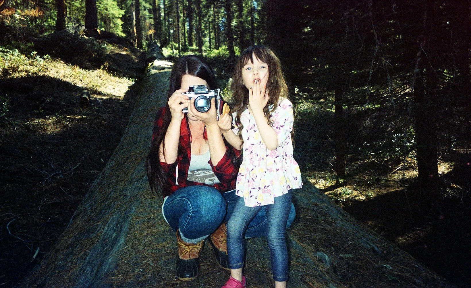 A woman with long dark hair, wearing a red plaid shirt and jeans, is sitting on a fallen tree in a forest and taking a photo with a camera. A young girl with curly brown hair, wearing a white floral shirt and denim jeans, is standing next to her, cov