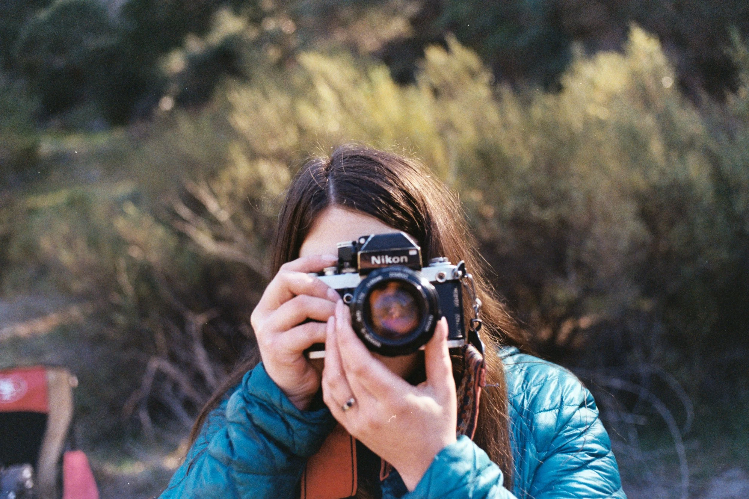 A person with long brown hair holding a Nikon camera, taking a photo outdoors with trees in the background.