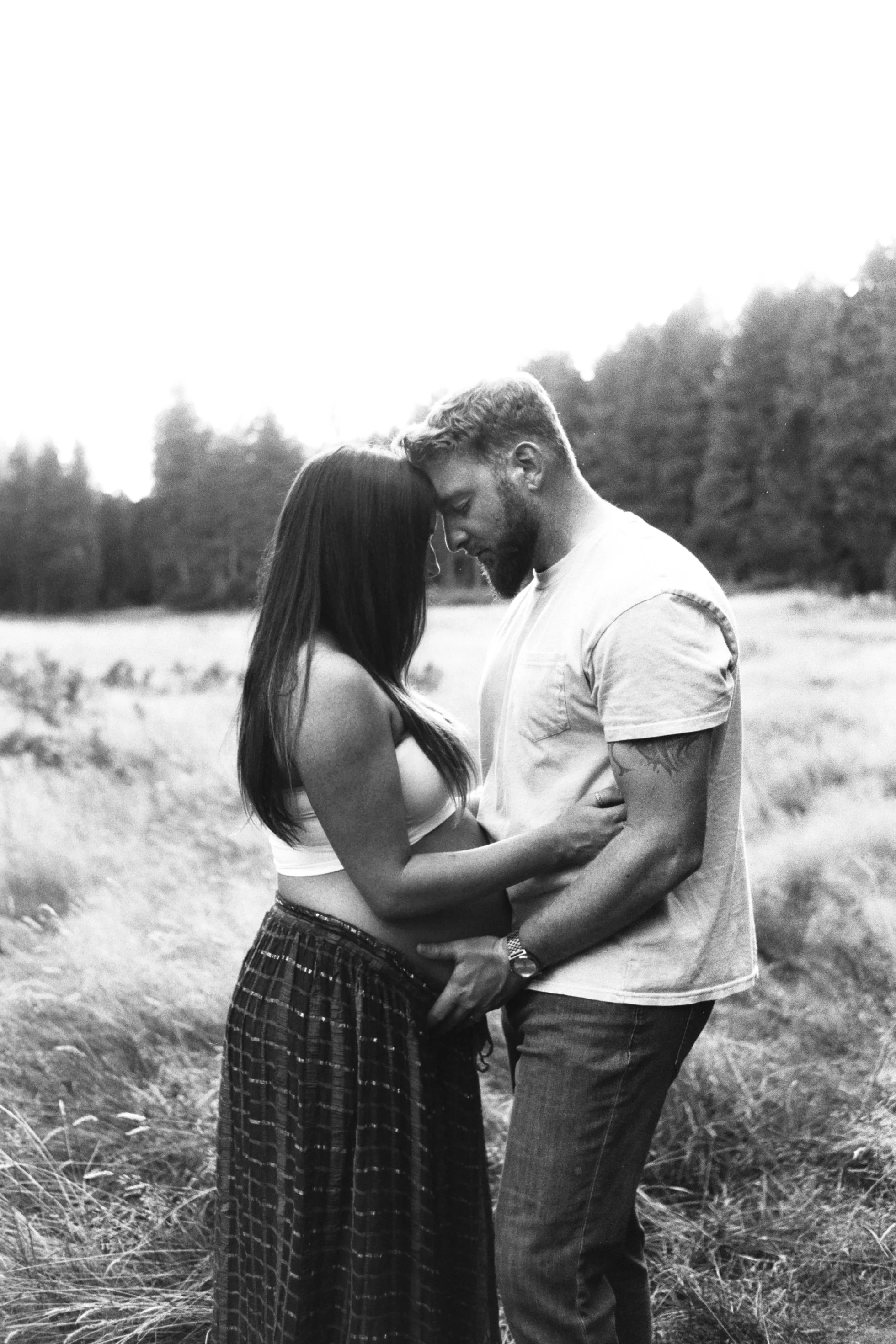 A black and white photograph of a couple standing close together in a field, their foreheads touching, with trees in the background.