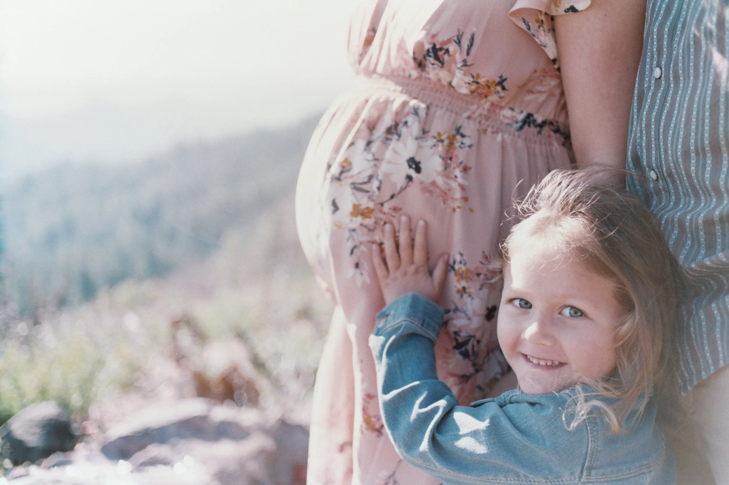 A woman and a young girl outdoors, smiling. The woman wears a pink floral dress, and the girl wears a denim jacket. The girl has blonde hair and blue eyes, and they are standing close together.