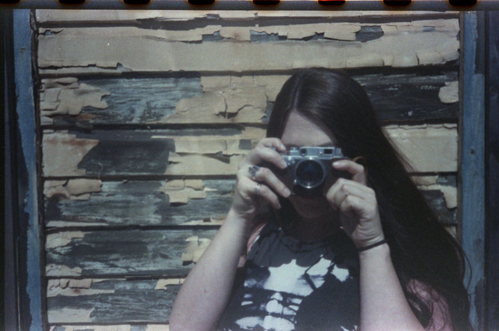 A woman taking a mirror selfie with a camera, standing in front of a weathered wooden wall with peeling paint.