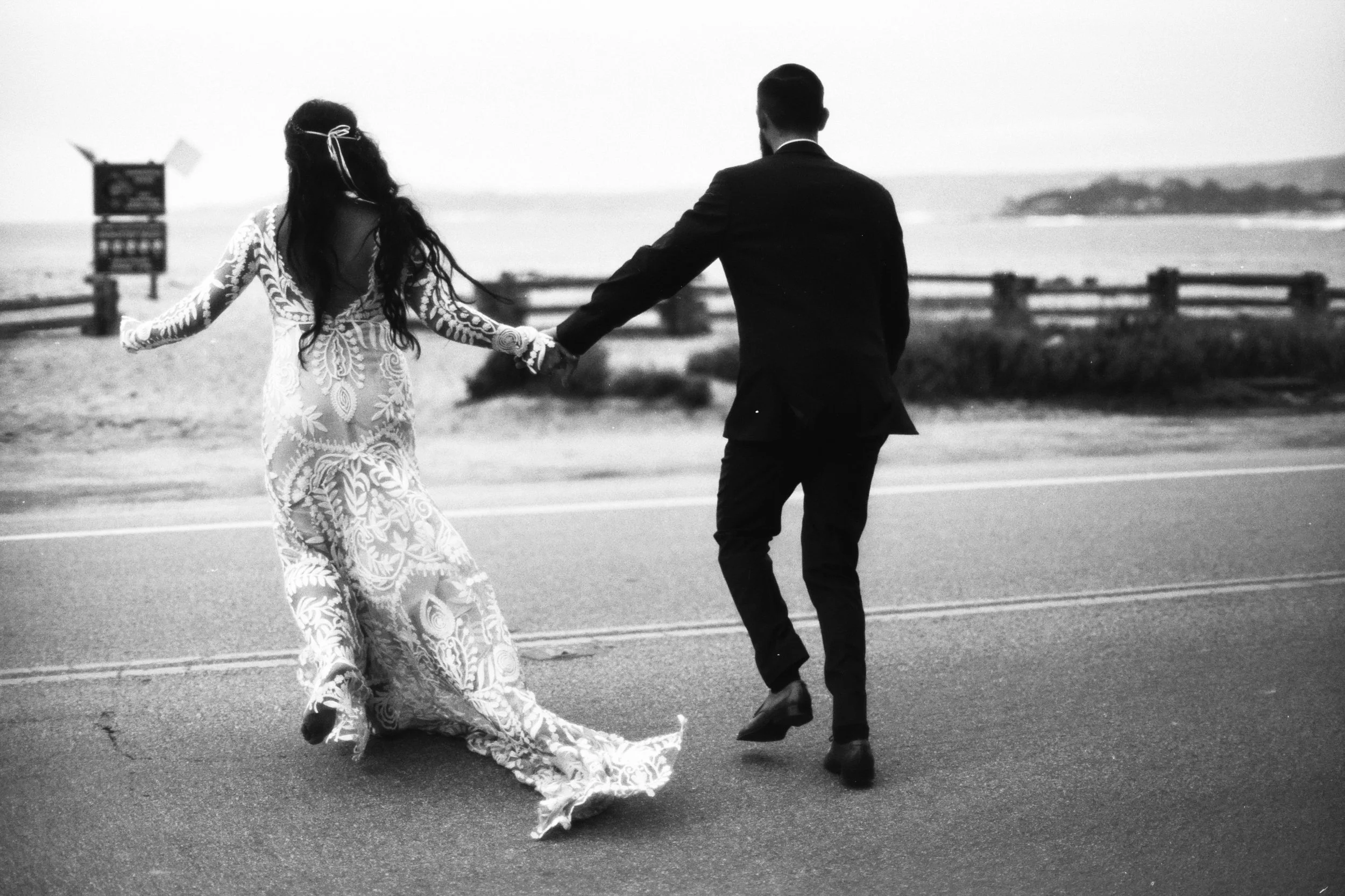 A black and white photo of a man and woman holding hands and dancing on a beach road. The woman wears a long, patterned dress and the man is dressed in a suit. The background shows the ocean and a distant landform.