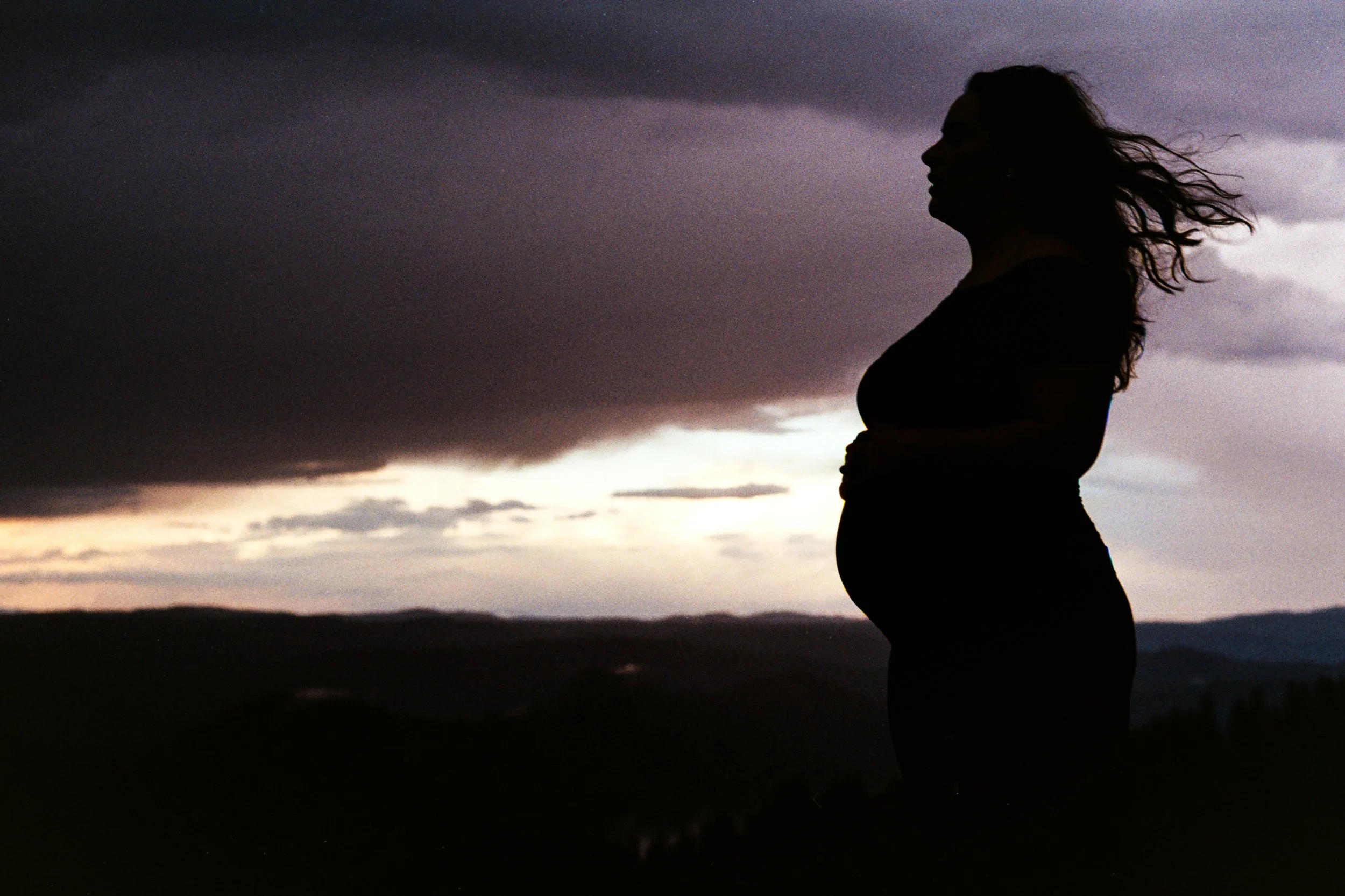 Silhouette of a pregnant woman standing outdoors during sunset, with dark clouds in the sky and distant landscape visible.