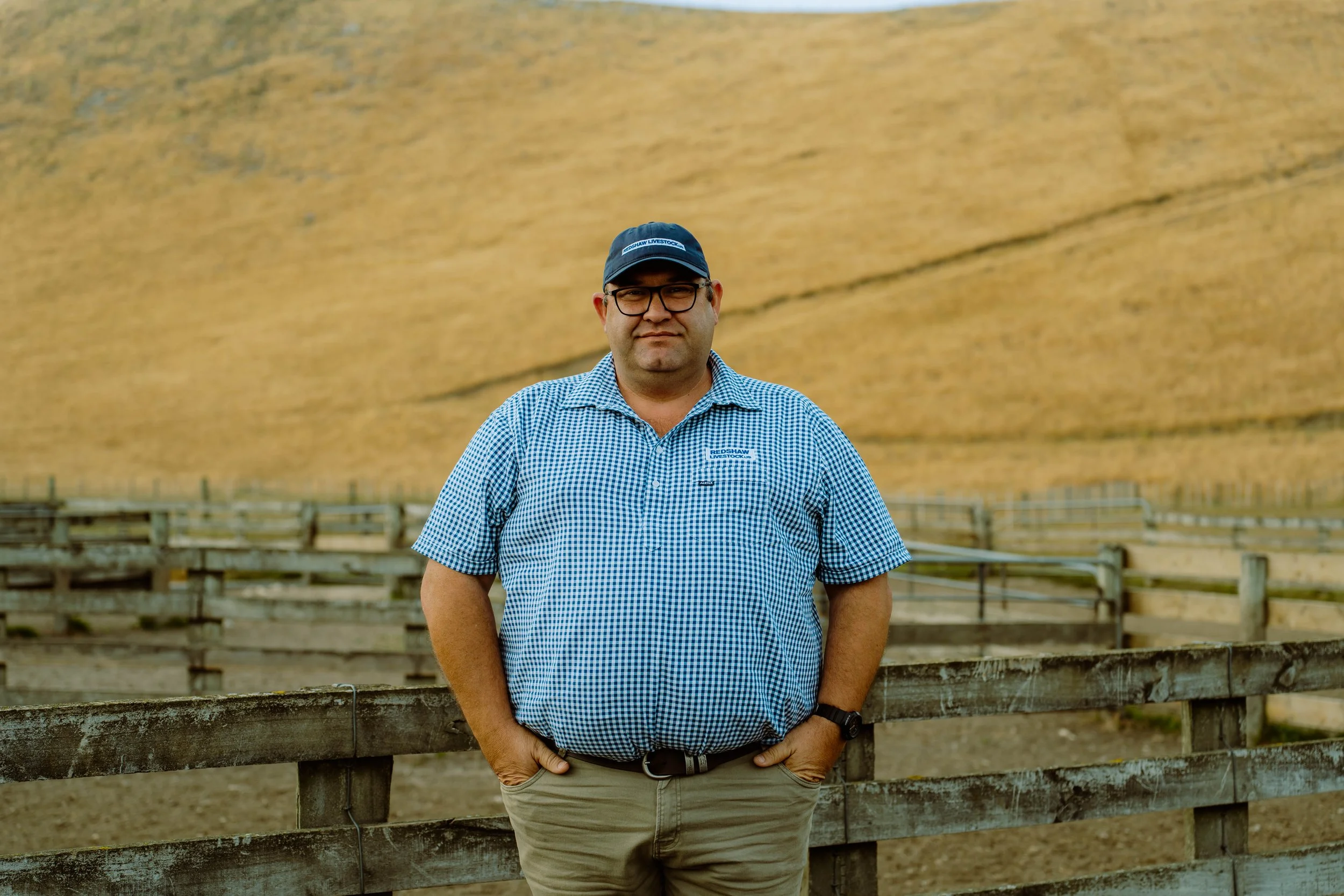 A man wearing glasses, a baseball cap, a blue checkered shirt, and khaki pants, standing outdoors near a wooden fence with a large haystack or mud wall in the background.