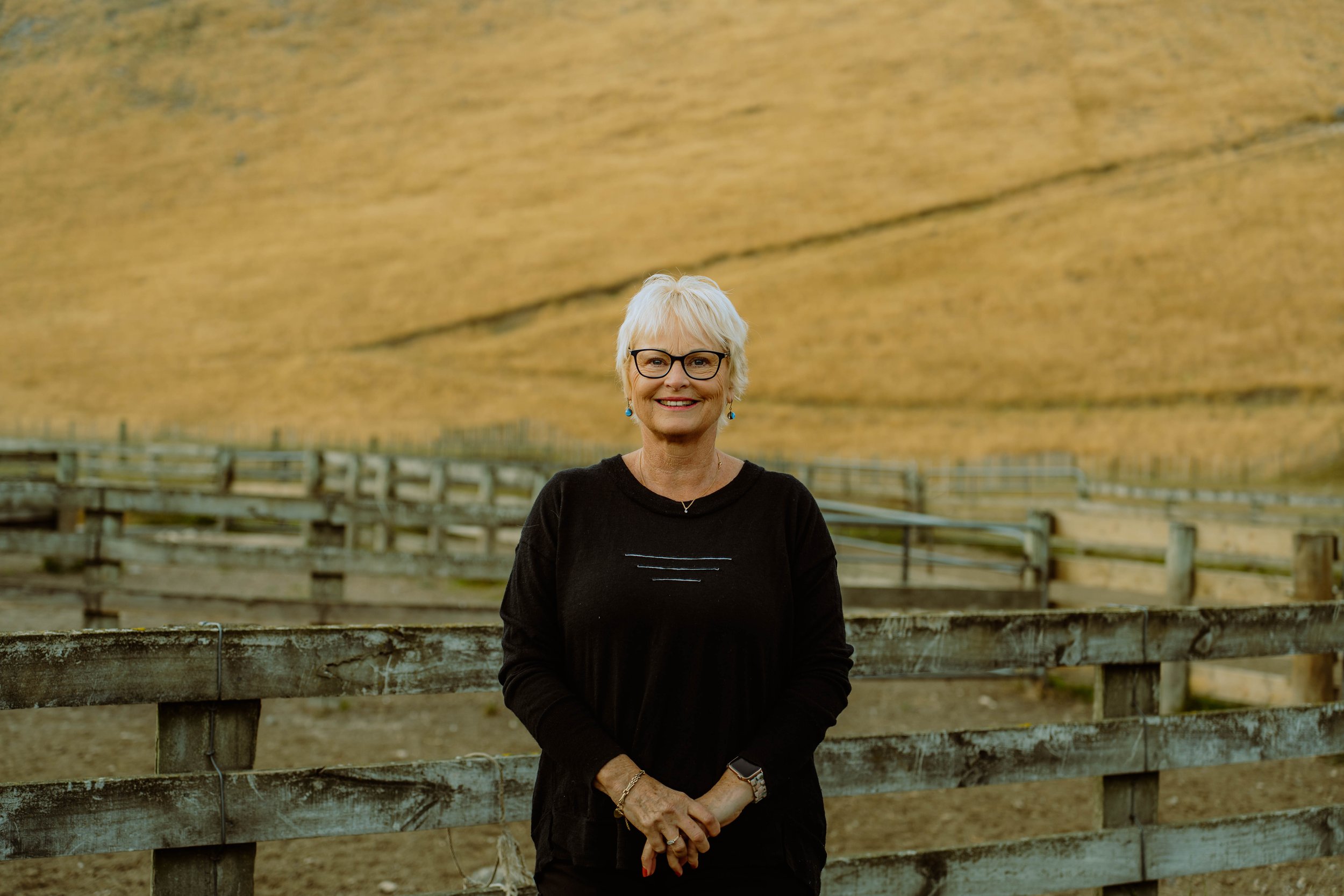 A smiling older woman with short white hair, glasses, and a black top, standing outdoors in front of a wooden fence with a large grassy hill in the background.