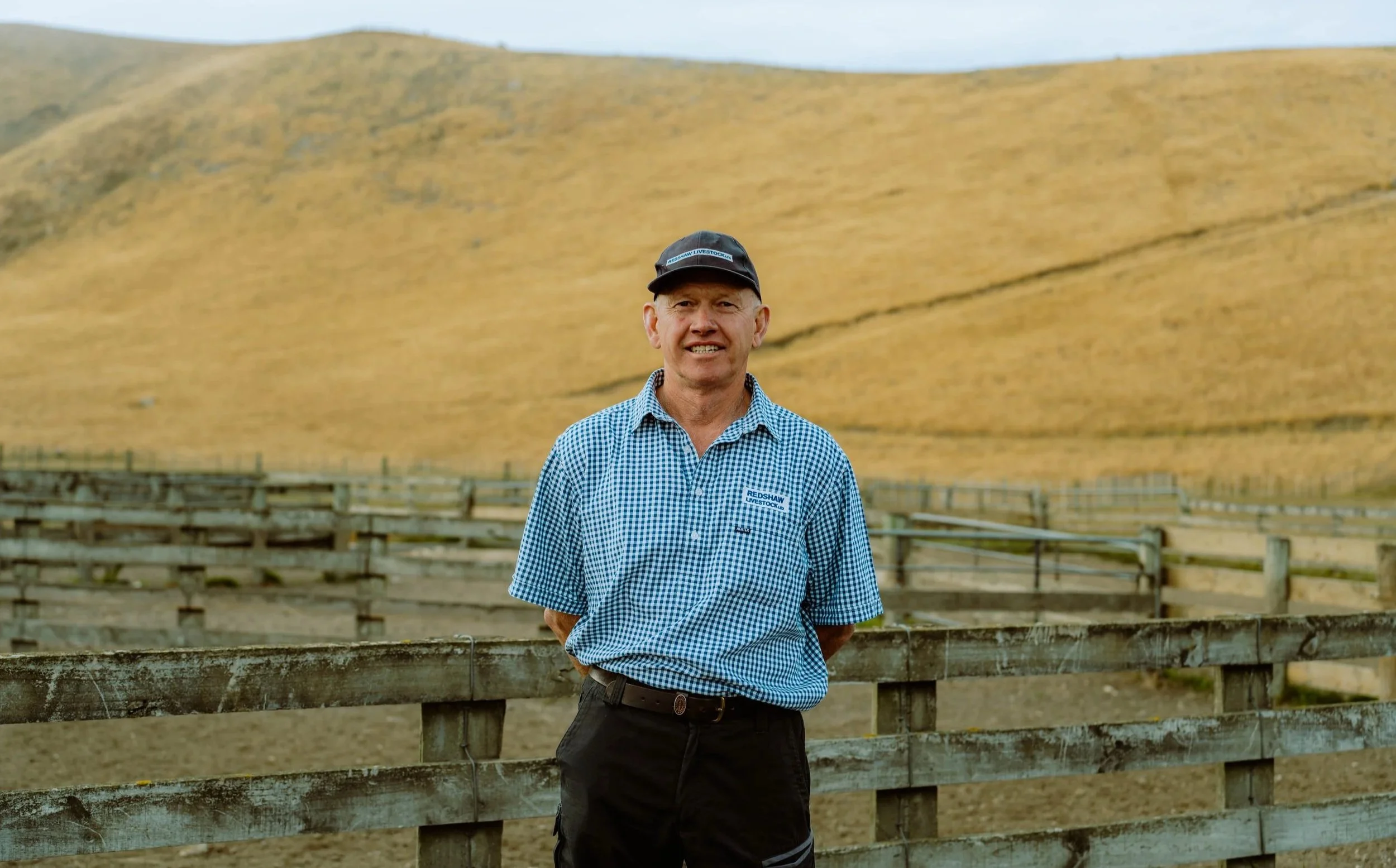A man wearing a checkered blue shirt and black cap standing outdoors in front of wooden fences and a large yellow grassy hill.