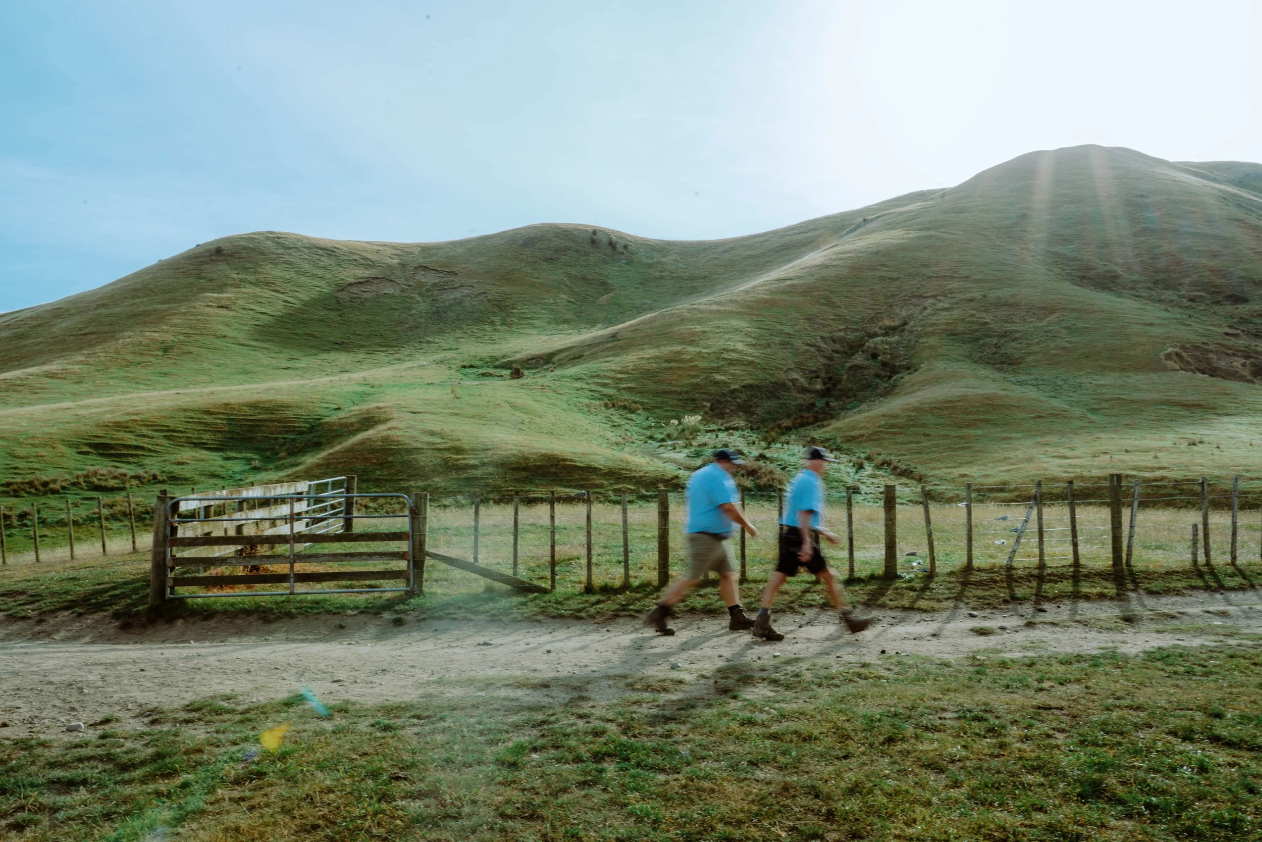 Two men walking past a fenced area with a backdrop of green rolling hills and a partly cloudy sky.