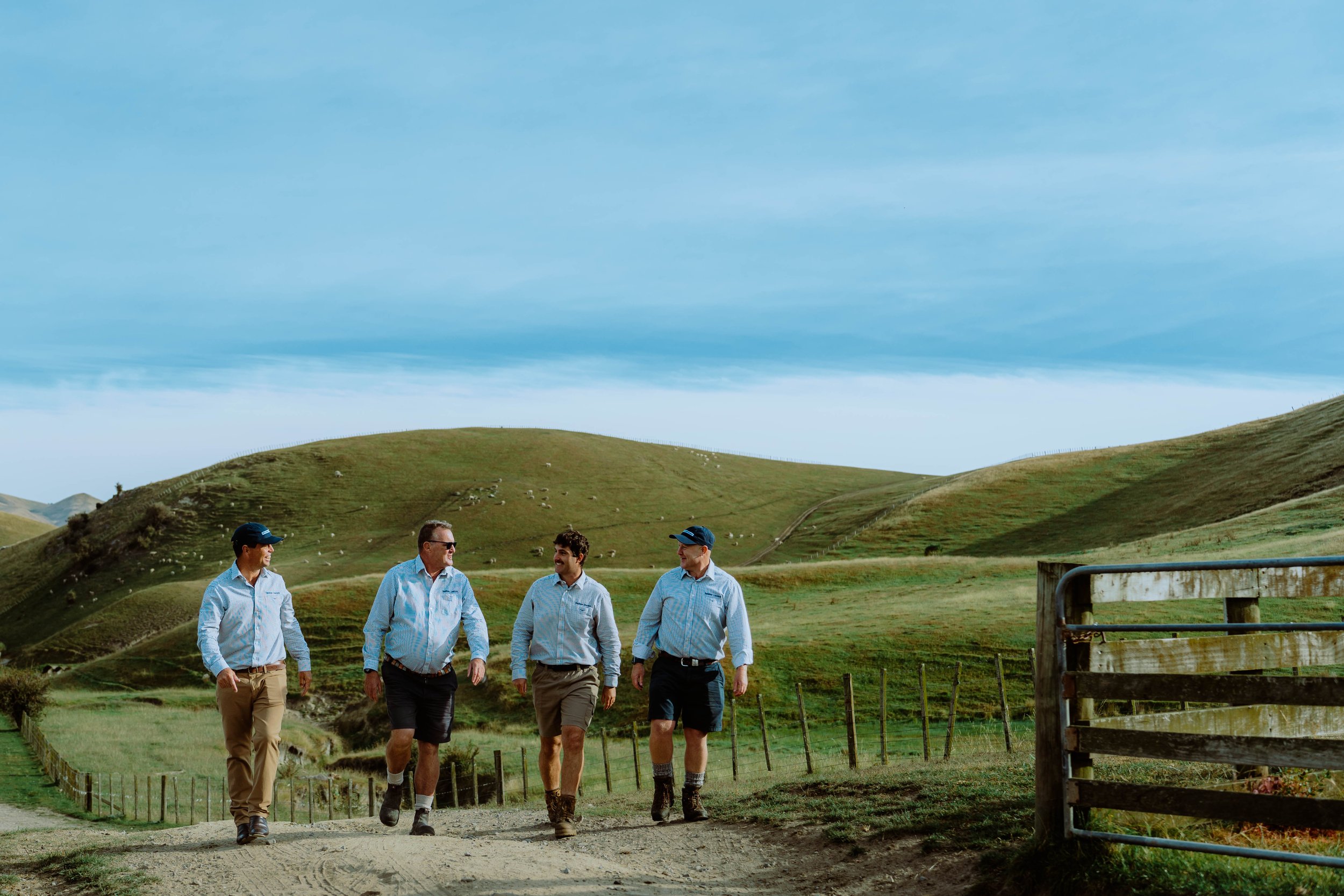 Four men in casual attire walking along a dirt path in a green hilly landscape under a blue sky with some clouds.