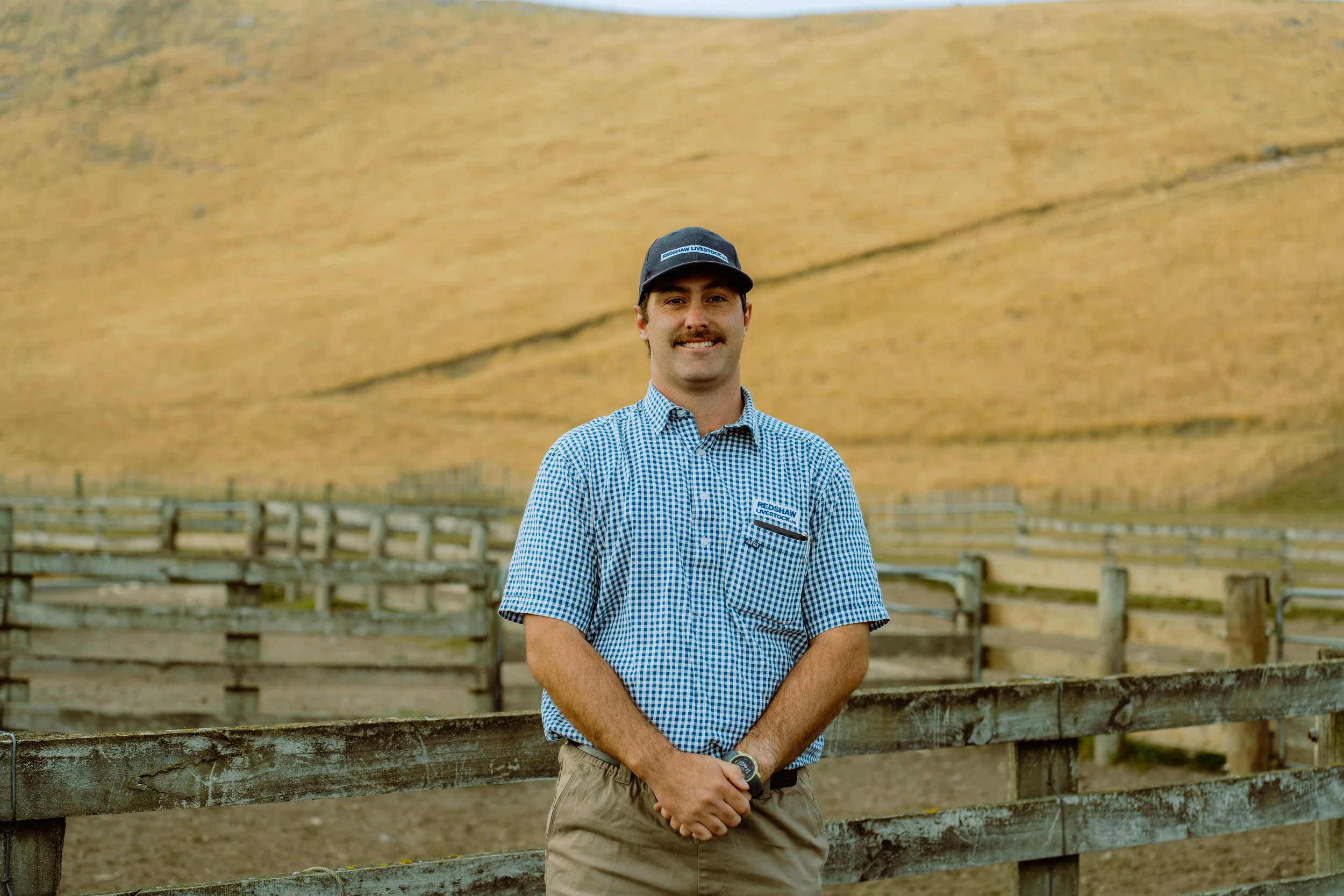 A man with a mustache and wearing a blue and white checkered shirt, a black cap, and khaki shorts stands outdoors in front of a wooden fence with large hay bales or silos in the background.