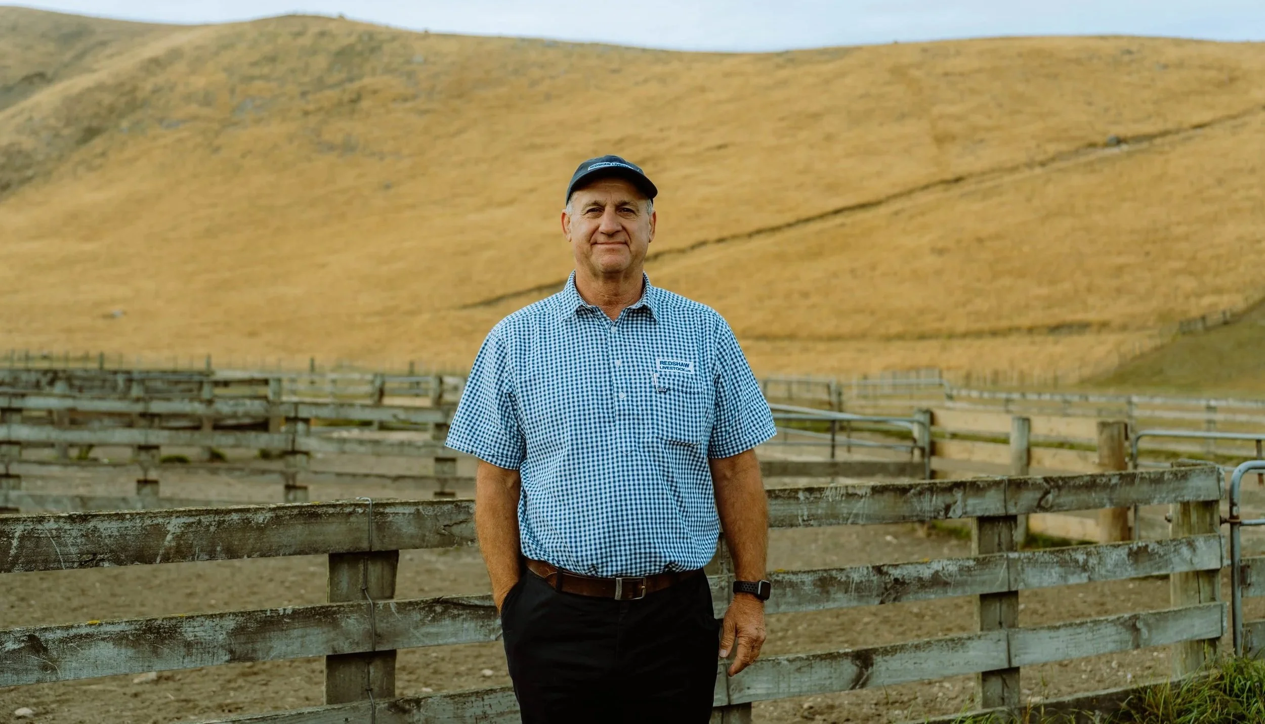 A man wearing a checkered shirt and a cap standing outdoors on a wooden fence with a large, golden hillside in the background.