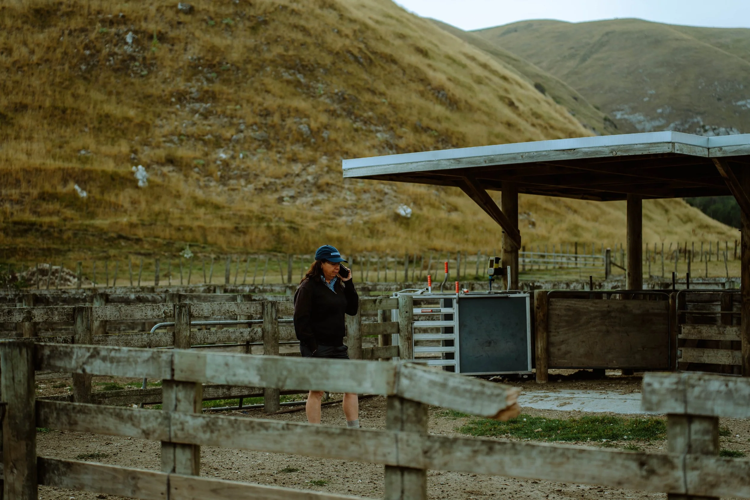 A woman in a black jacket and blue hat standing near a wooden fence at an outdoor farm or ranch, with a green and brown hillside in the background. She is talking on a cellphone and surrounded by farm equipment.