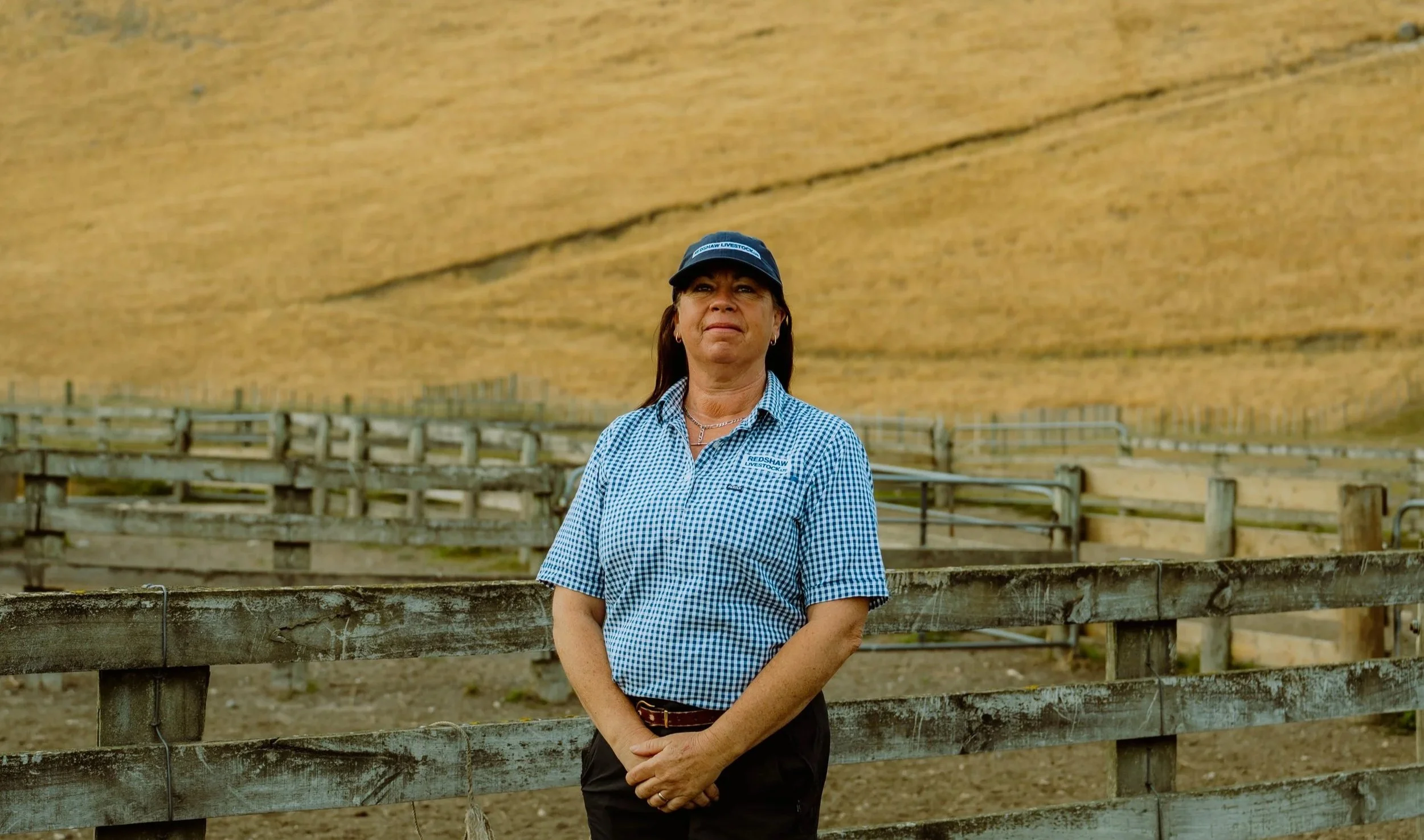 A woman standing near a wooden fence outdoors, wearing a blue checkered shirt, a black cap, and a necklace, with a sunny weather and a large hill or mountain in the background.
