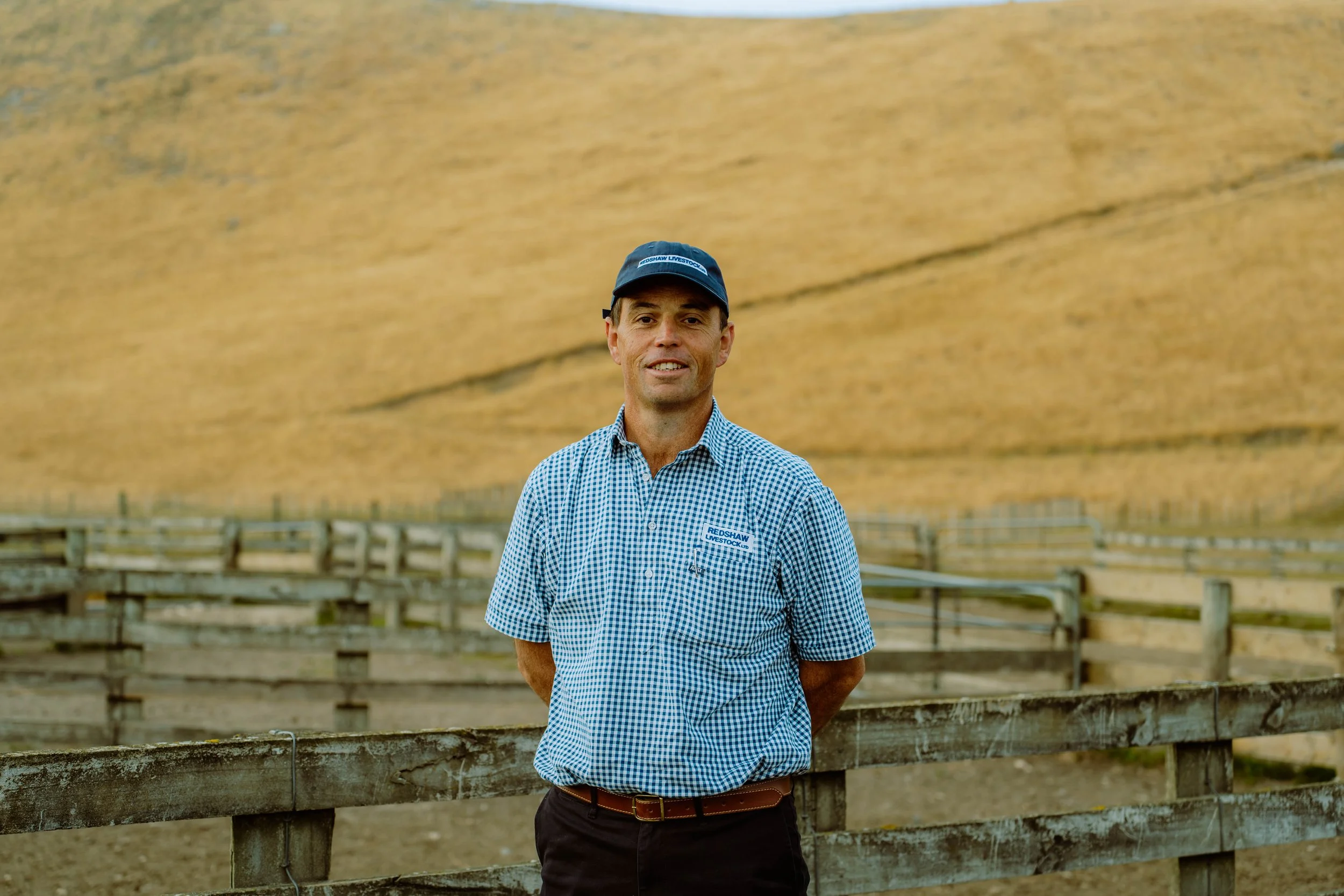 A man wearing a blue checkered shirt and a cap stands outdoors in front of a wooden fence, with a large, golden-brown straw or haystack in the background.