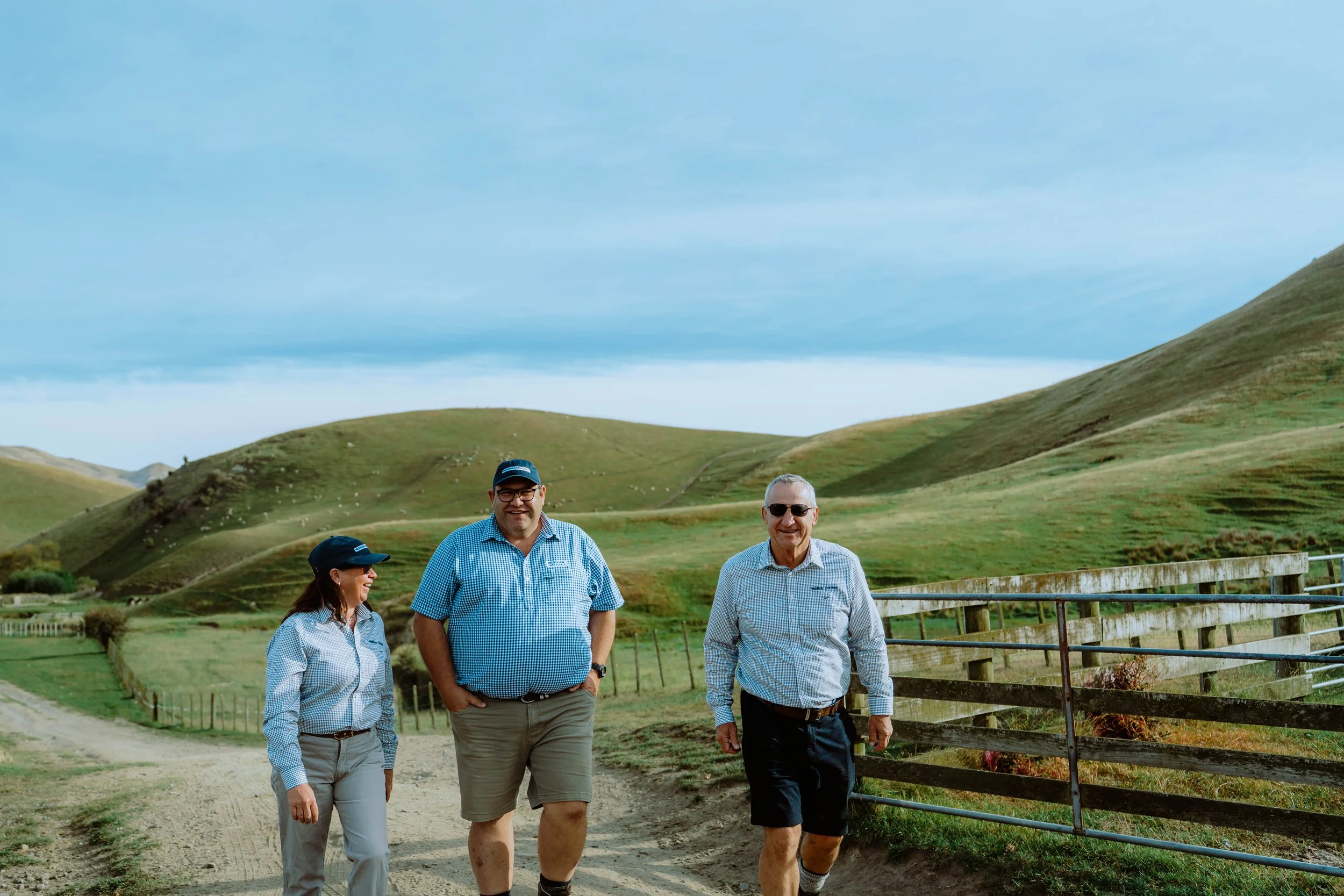 Four livestock agents walking outdoors on a dirt path with green rolling hills and a cloudy sky in the background. Two men and one woman are visible, all smiling and dressed in casual outdoor clothing, including hats and sunglasses.