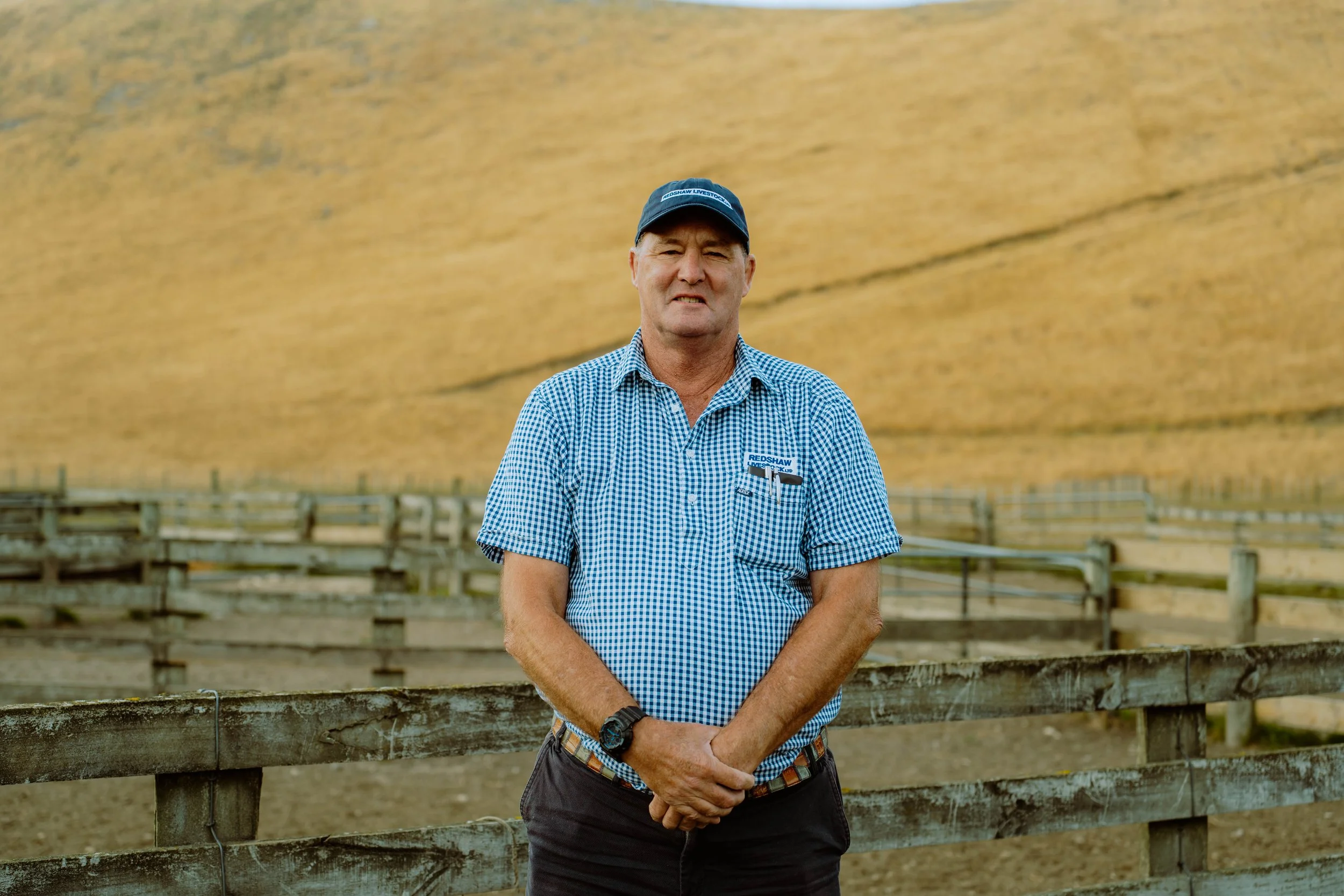 A man standing outdoors near a fenced area, wearing a blue checked shirt, a black cap, and a watch, with a large yellowish hill or wall in the background.