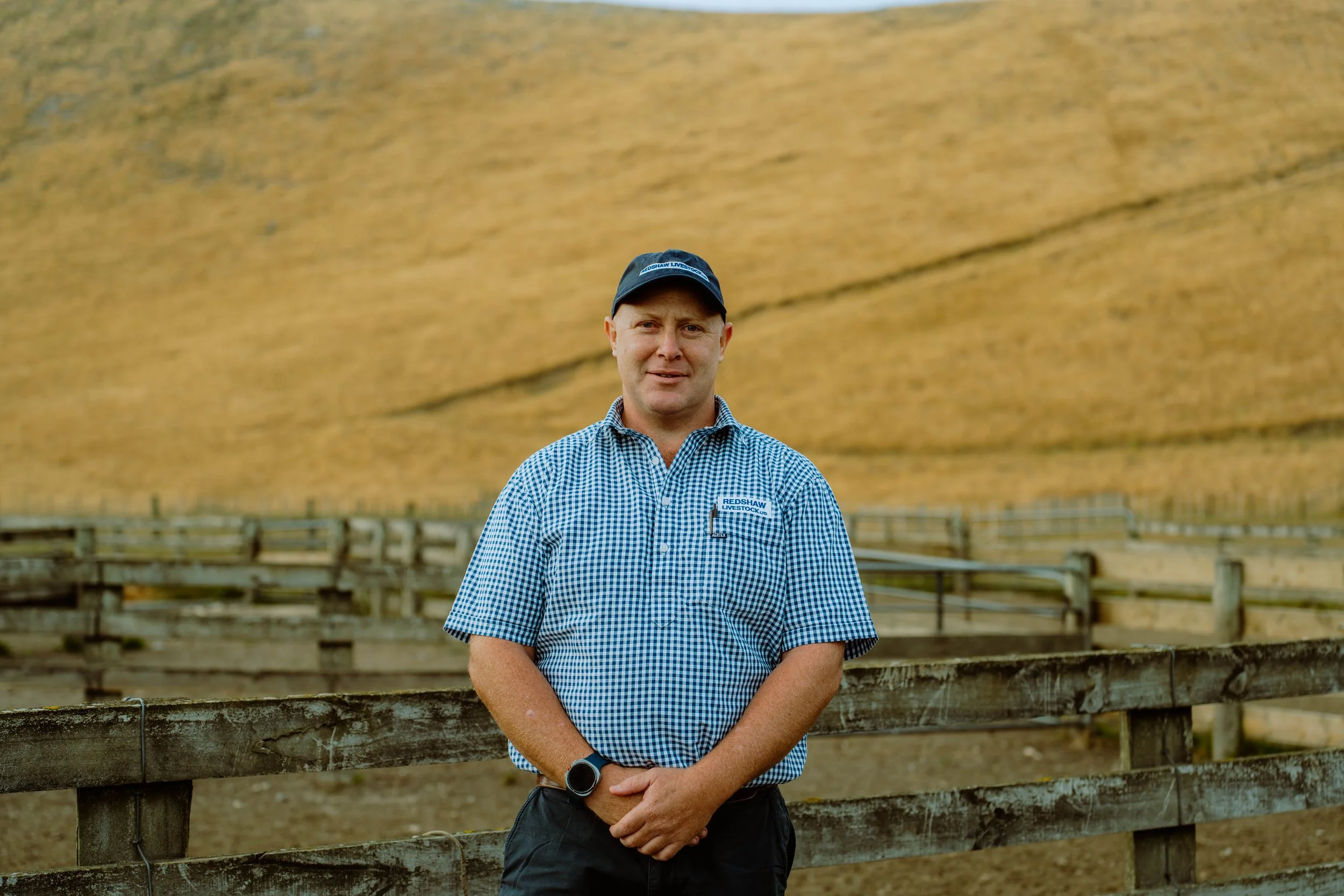 A man in a checkered blue shirt and blue cap standing outdoors in front of a fenced pasture with yellowish-brown hills in the background.