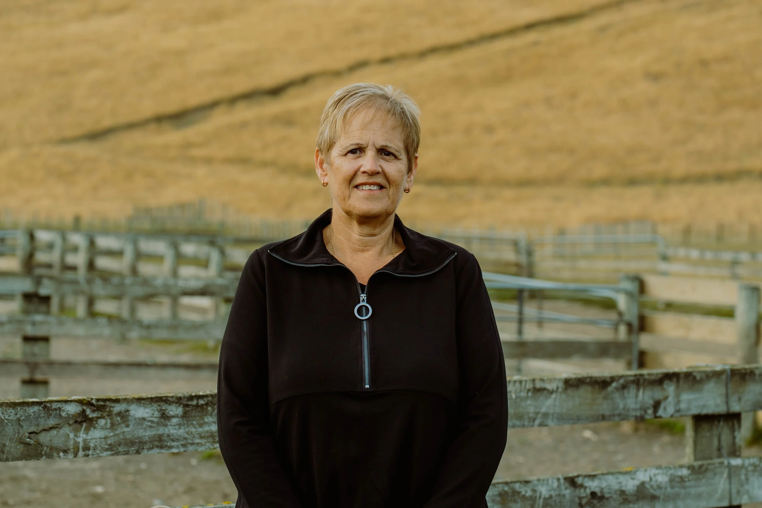 A middle-aged woman with short blond hair wearing a black zip-up jacket, standing outdoors in front of a wooden fence with a deserted landscape background.