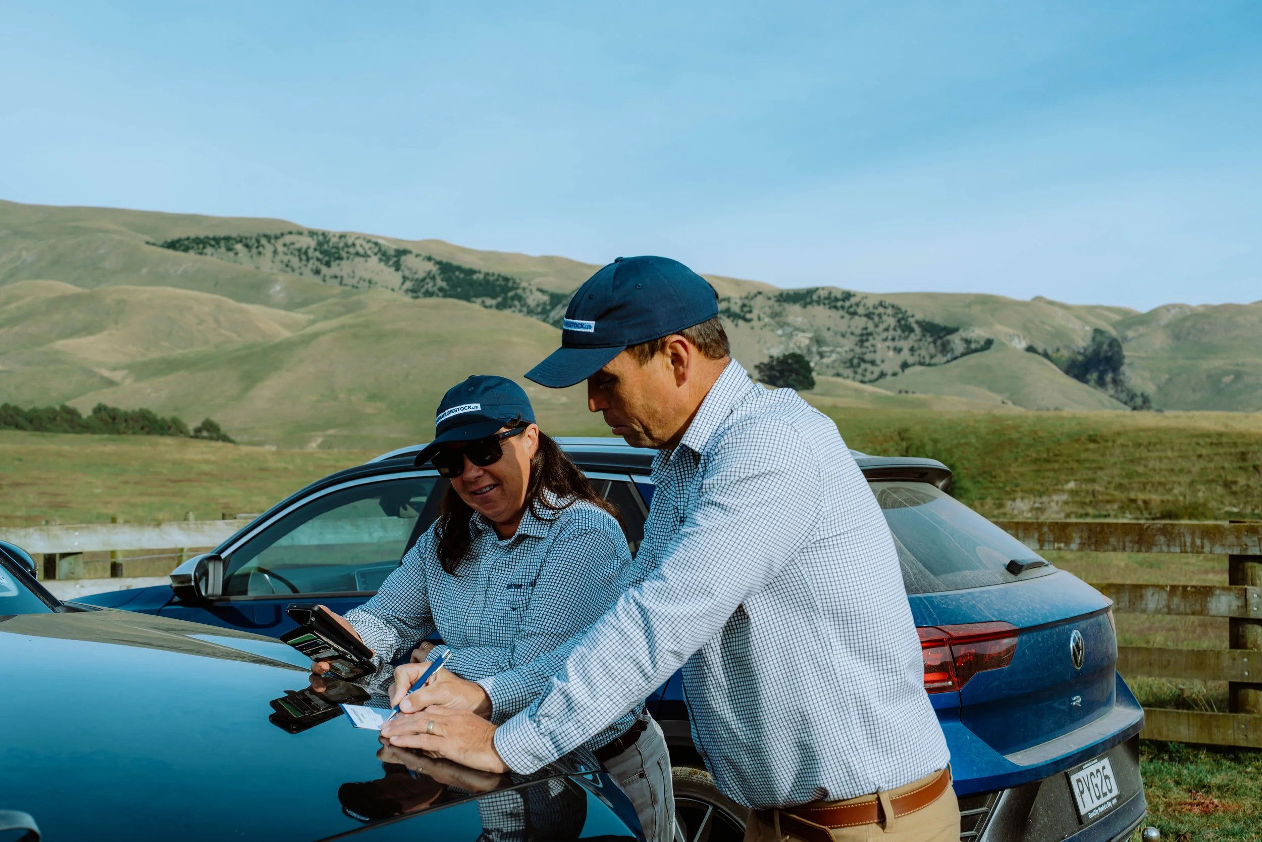Two people wearing blue caps and checkered shirts standing next to a blue car, outdoors in a hilly area, looking at a handheld device.