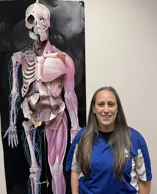 A woman standing in front of a poster of a human anatomy model showing muscles and bones.