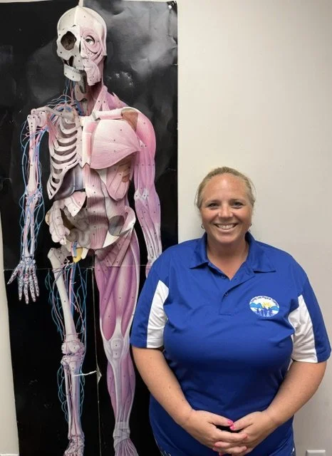 A smiling woman standing next to a large anatomical poster of the human body, showing muscles, bones, and nerves.