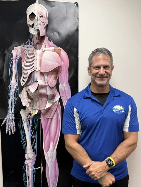 A man smiling and standing next to a human anatomical model showing muscles, bones, and nerves.