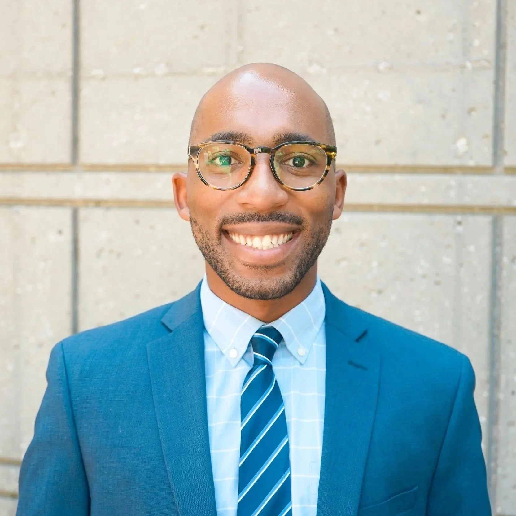 A smiling man in a blue suit, light blue shirt, and striped tie standing against a light-colored brick wall, wearing glasses with tortoiseshell frames.
