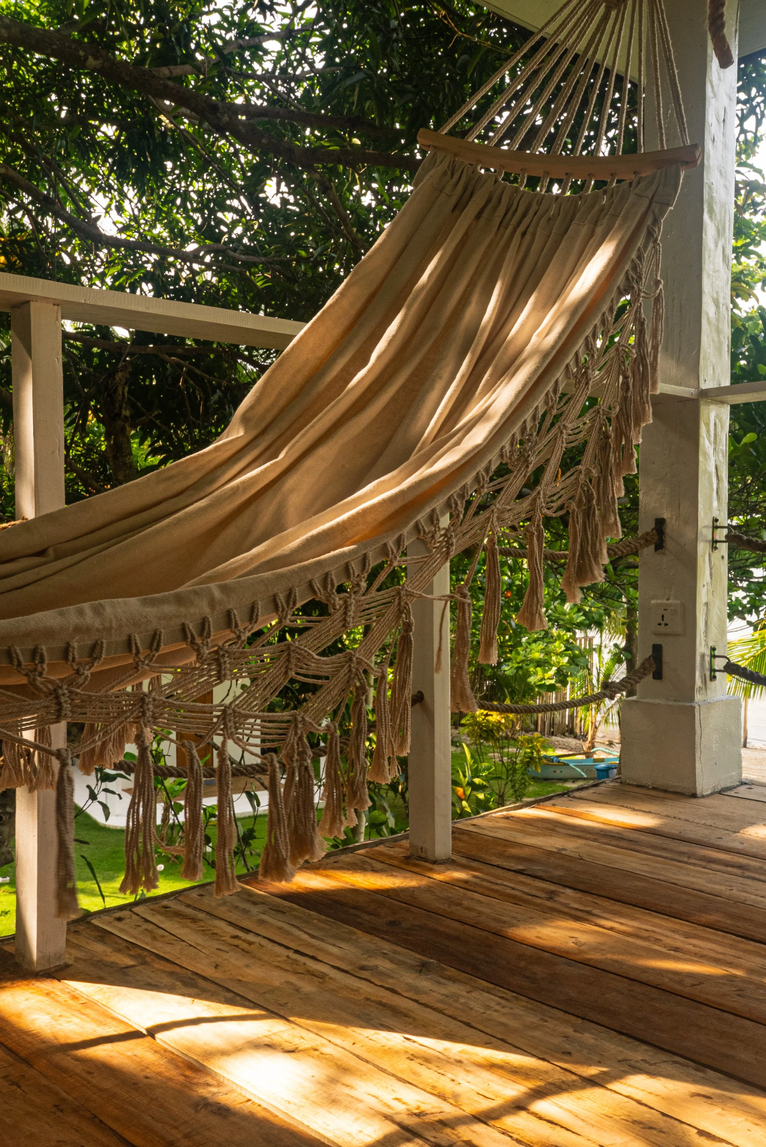 Un hamac beige attaché à une structure en bois sur une terrasse en bois, entouré de végétation et de lumière naturelle.