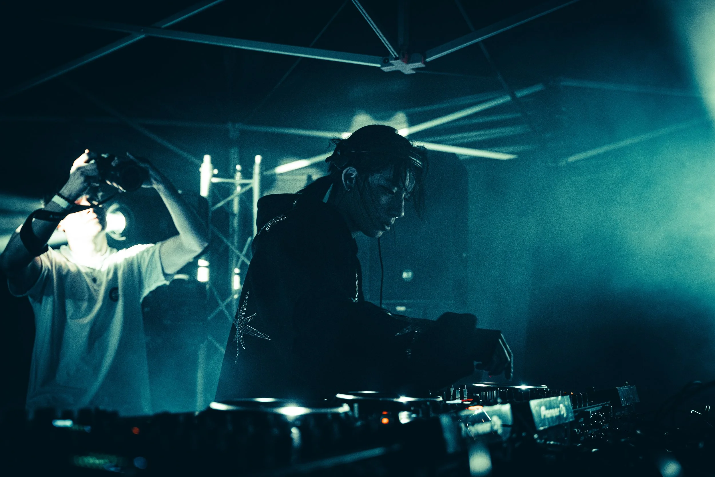 DJ jouant de la musique dans une boîte de nuit sombre avec des lumières colorées et des spectateurs en arrière-plan.